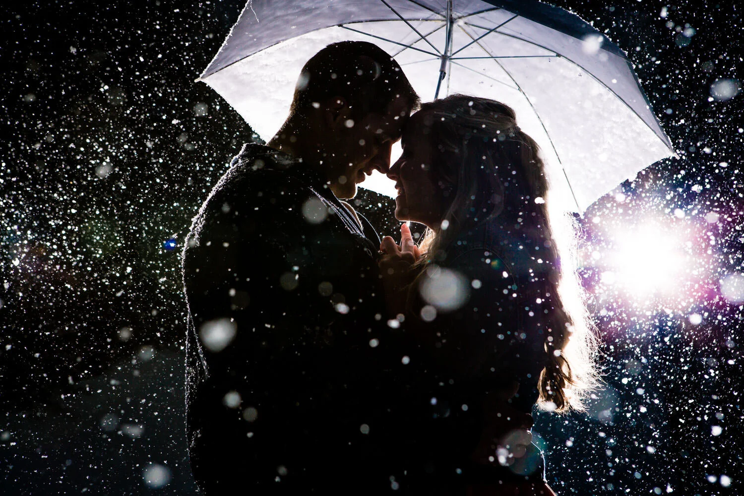 Silhouetted couple under an umbrella in the rain at night, with light shining behind them.