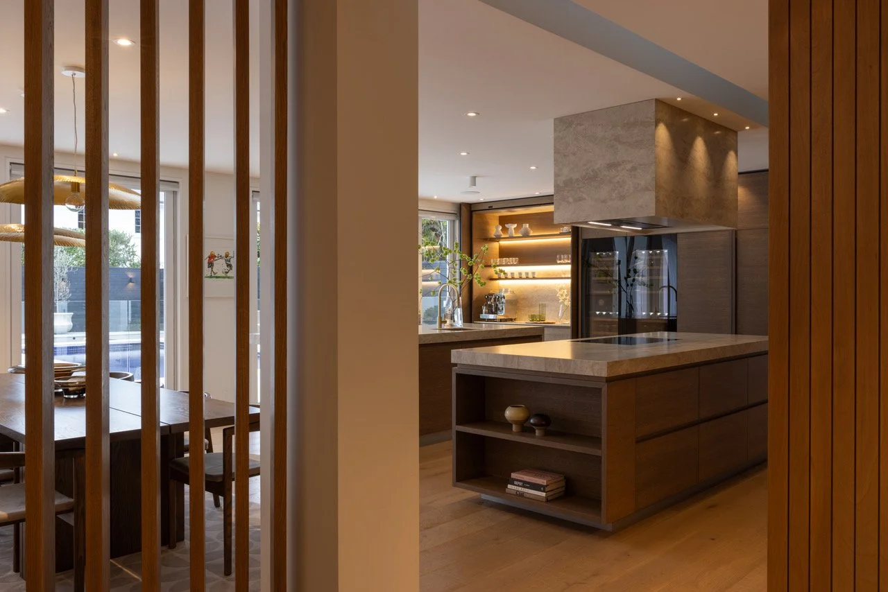 Modern kitchen with island, built-in shelves, and warm lighting, viewed through wooden room divider by Eternodesign