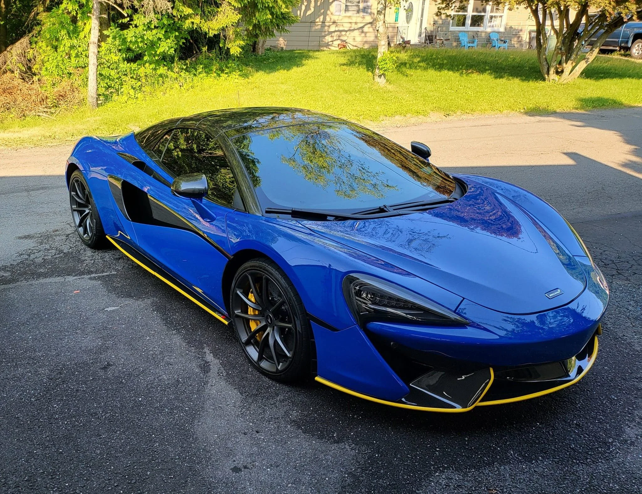 A blue sports car with black details and yellow accents parked on a wet asphalt road in a residential area, with trees and grass in the background.