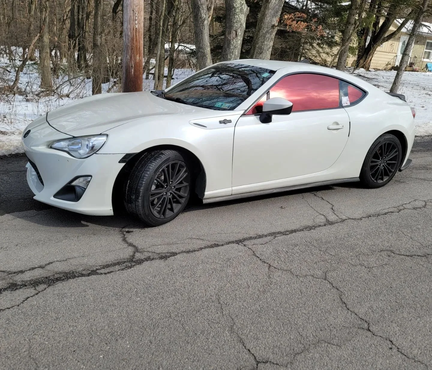 White coupe car parked on a cracked asphalt road with snow and trees in the background.