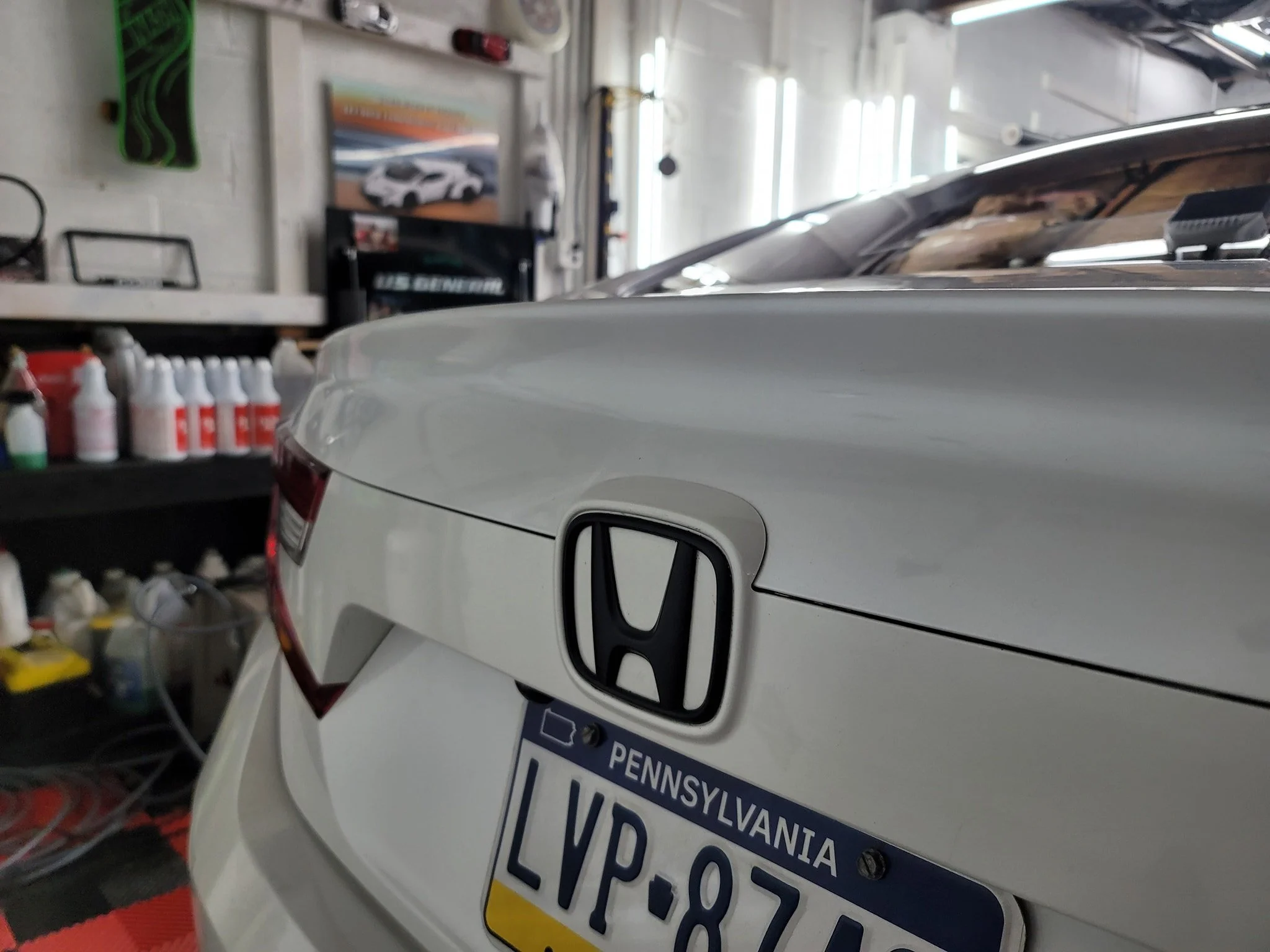Rear of a white Honda car with Pennsylvania license plate, inside a workshop with shelves holding bottles of car products and tools, some posters on the wall, and fluorescent lights overhead.