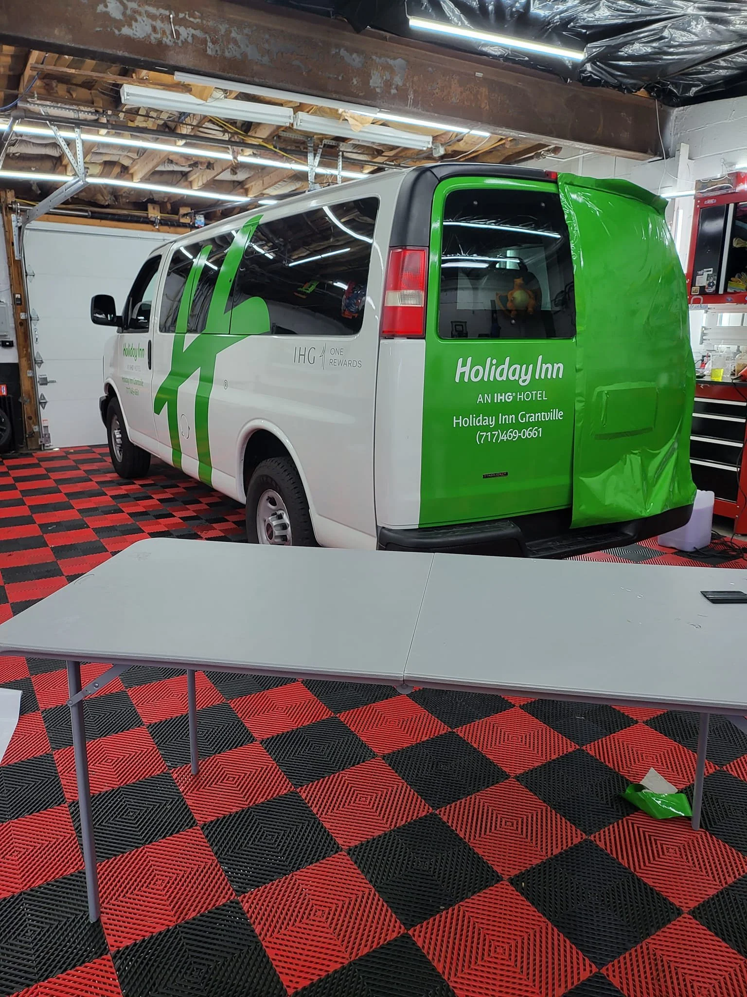 A Holiday Inn branded van inside a garage with red and black checkered flooring, a folding white table in the foreground, and garage tools and equipment on the sides.