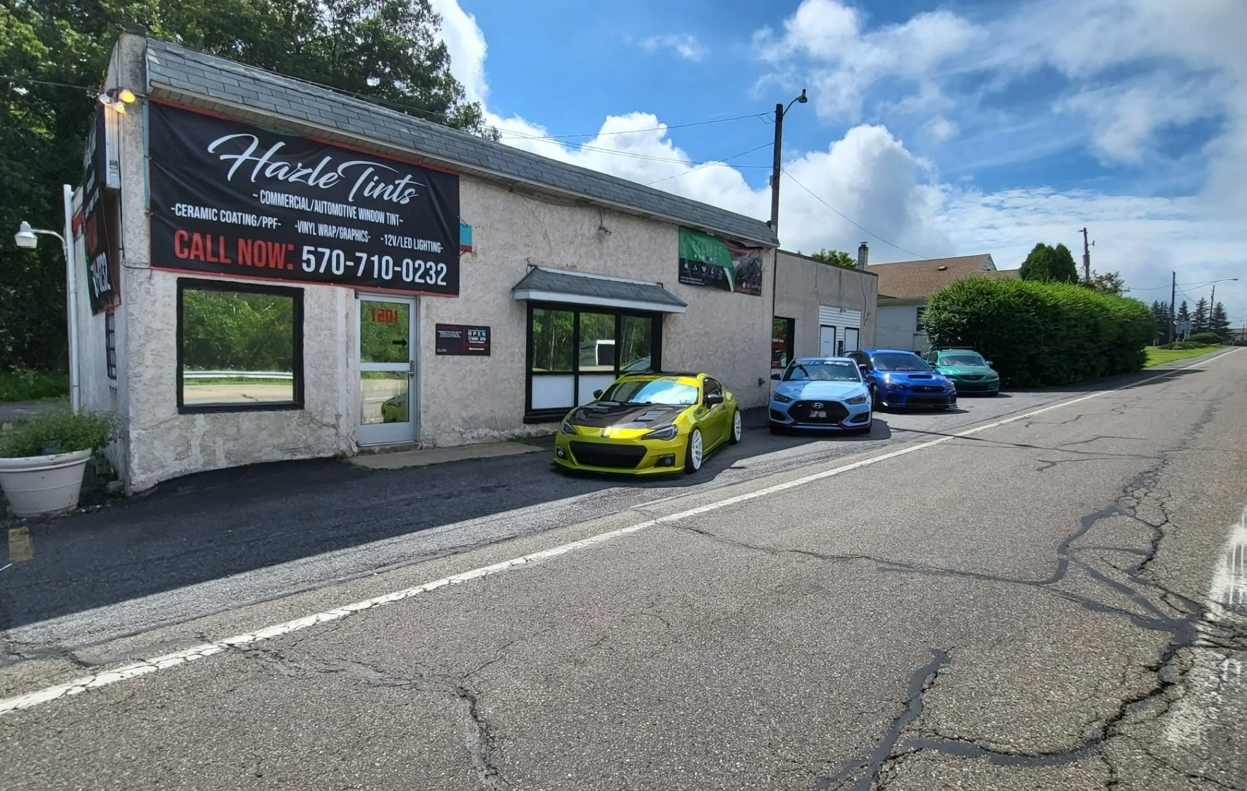 Auto repair shop with a sign that reads 'Hale Tints' and a phone number, with four cars parked in front: a yellow sports car, a blue sedan, a black coupe, and a green hatchback, on a sunny day.