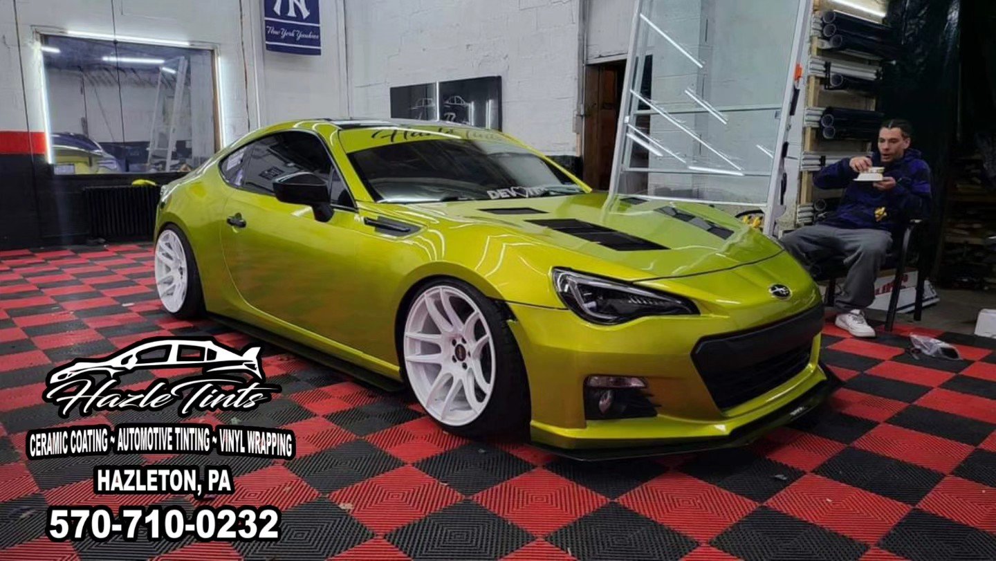 A green sports car with black accents inside a garage or workshop, next to a man sitting on a chair eating. The garage has a red and black checkered floor, and branding for Hazle Tints, an automotive tinting and wrapping business, is visible on the i