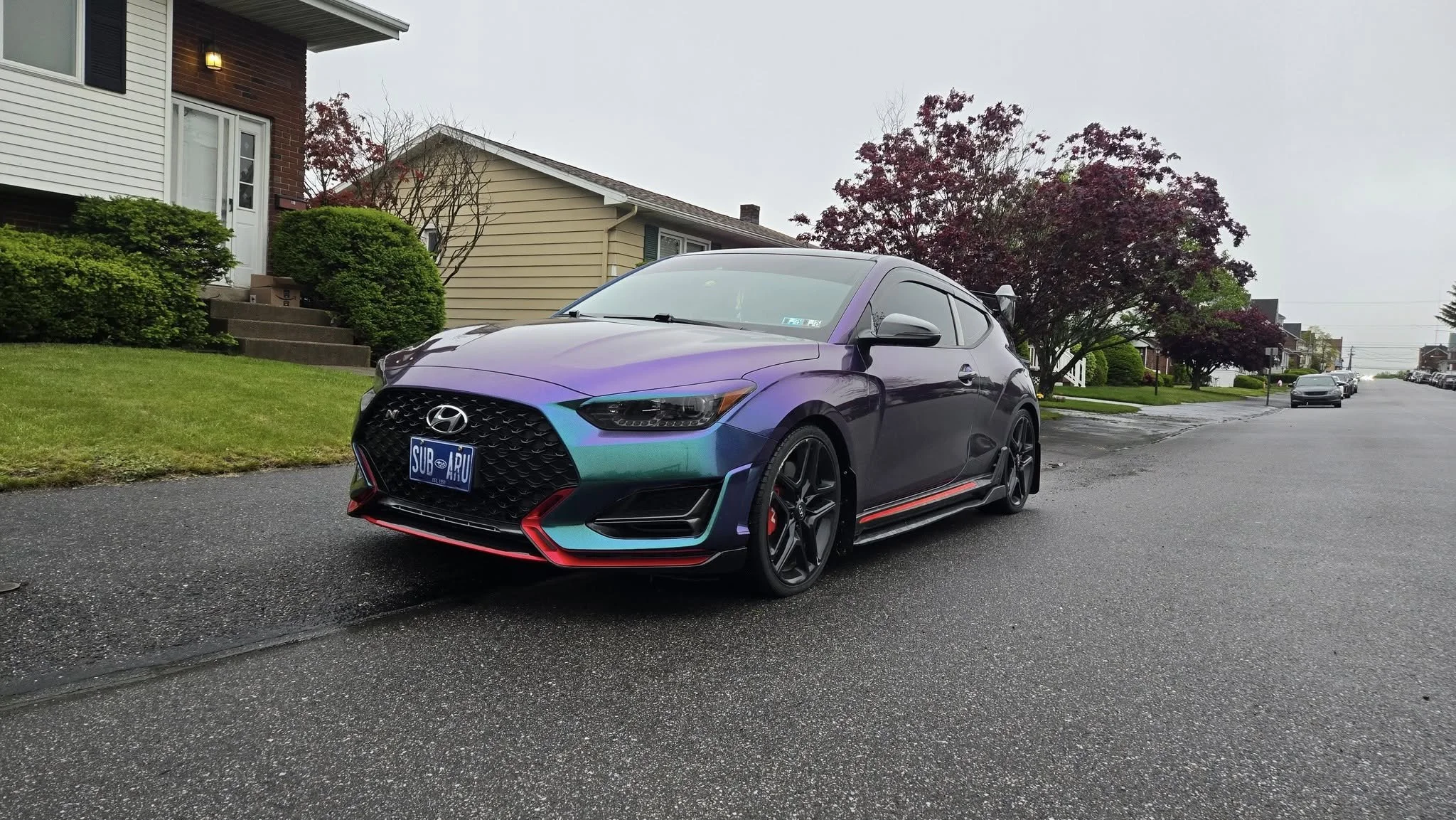 A metallic purple and blue Hyundai sports car parked on a suburban street with houses, trees, and other cars in the background.