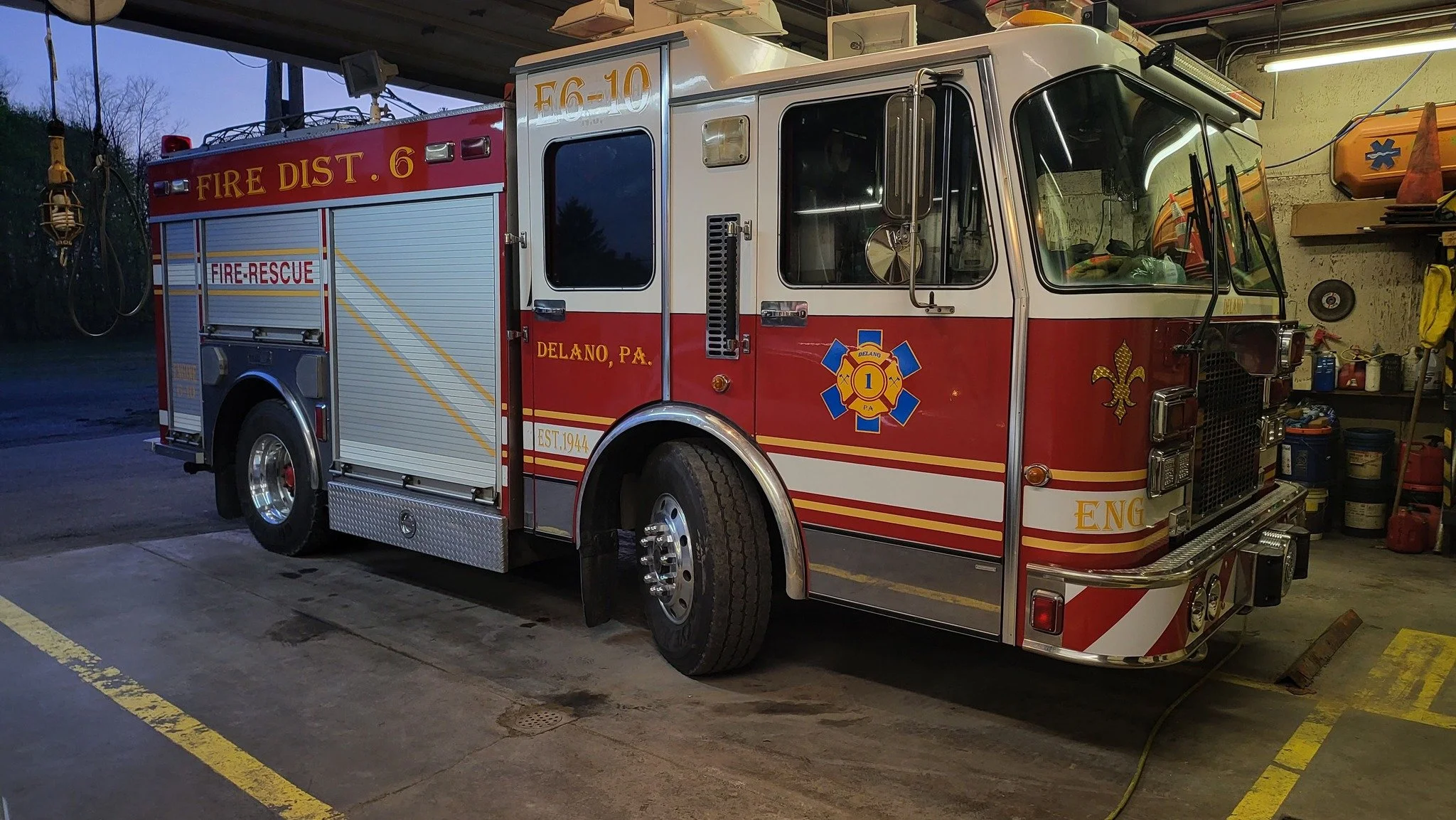 A fire rescue truck parked inside a fire station garage. The truck has red and white colors, with emergency lights on top, and the words 'FIRE DIST. 6' and 'DELANO, PA' on the side. There are various tools and equipment on the wall behind the truck.