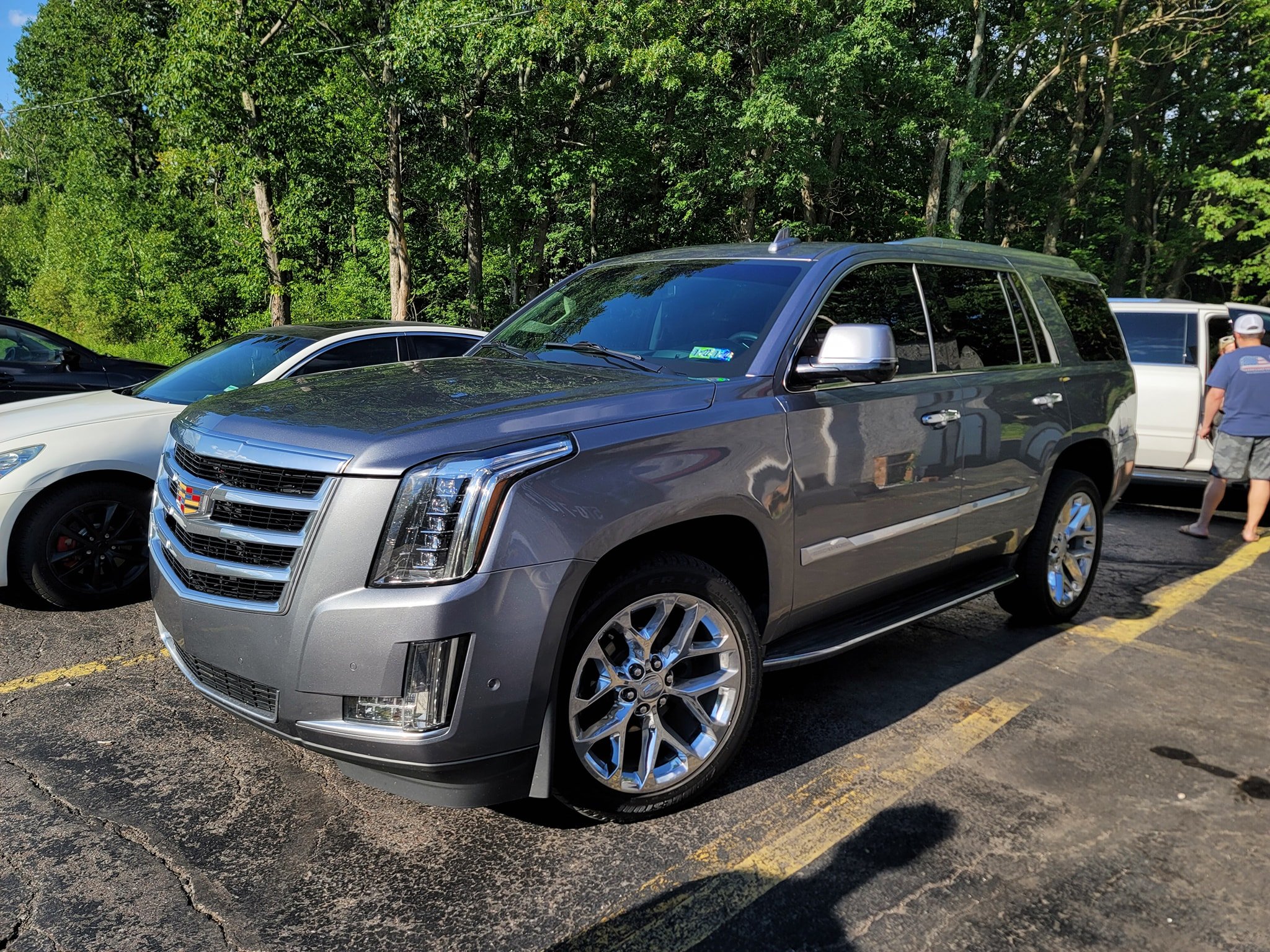A gray Cadillac SUV parked in a parking lot with trees in the background and a person walking past it.