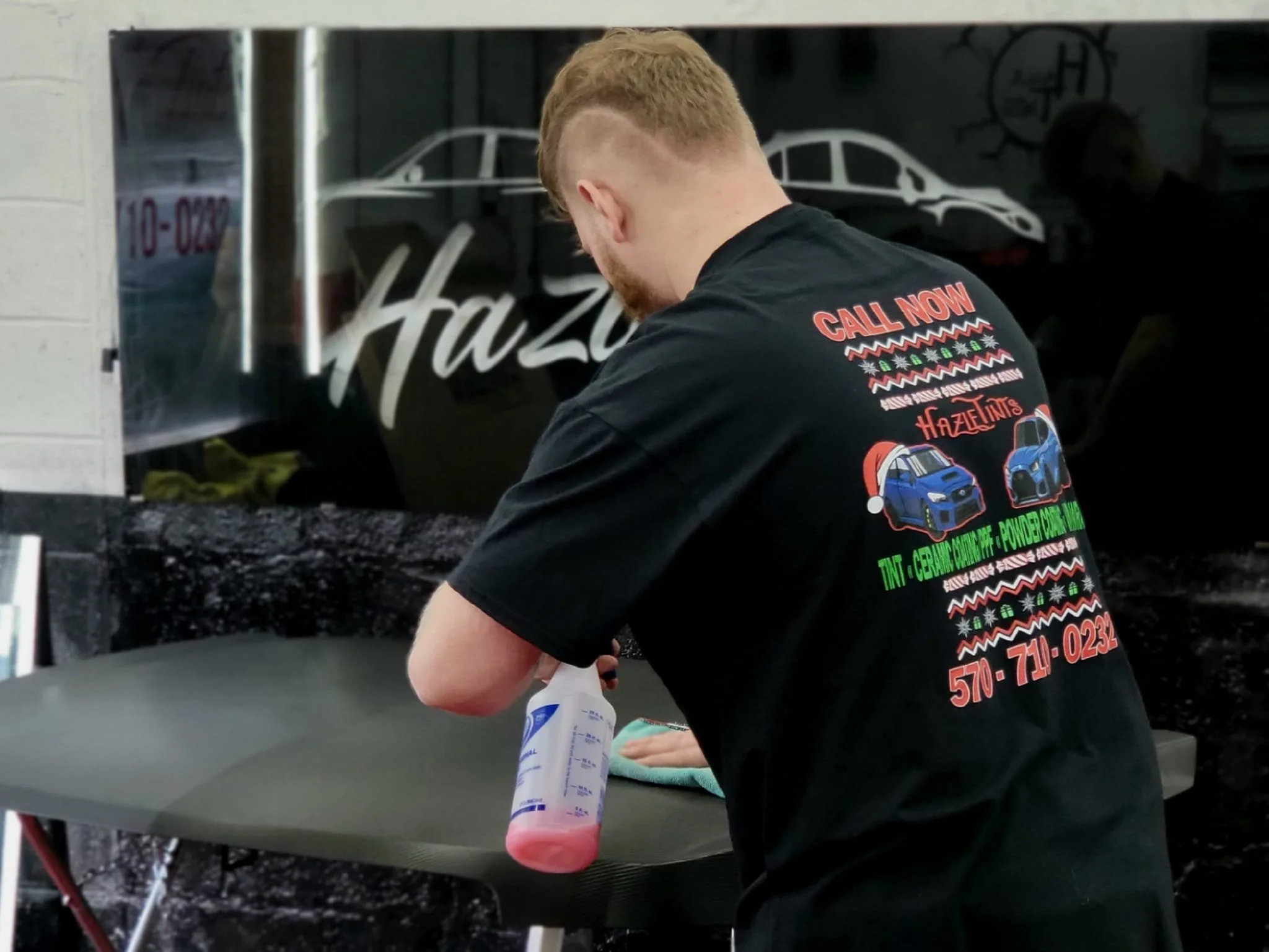 A man in a black T-shirt working with a spray bottle and cleaning cloth at a table, with a glossy black background that has white graphic design of cars.