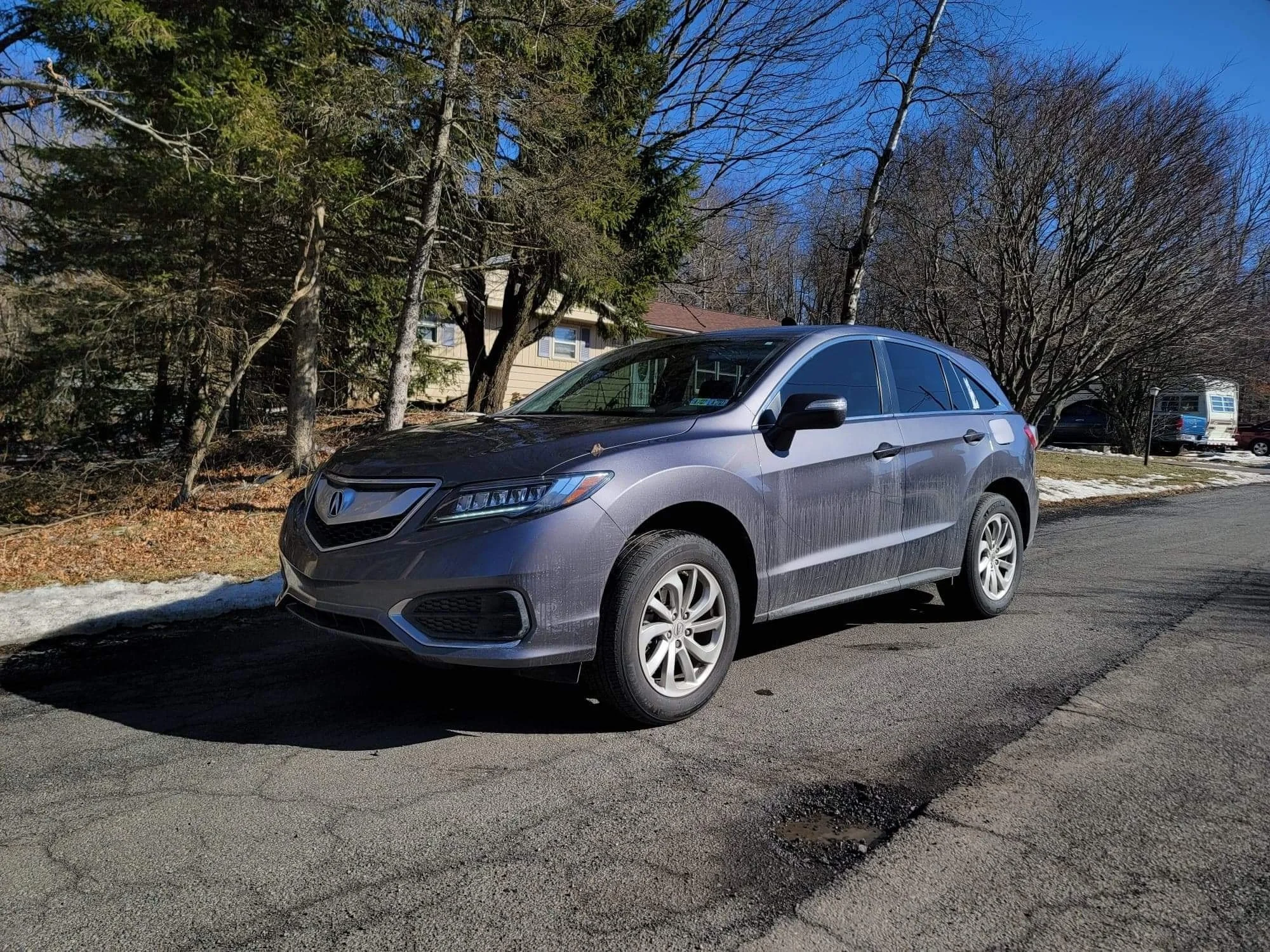 A grey Acura SUV parked on a street with leafless trees, a house, and other vehicles in the background on a clear winter day.