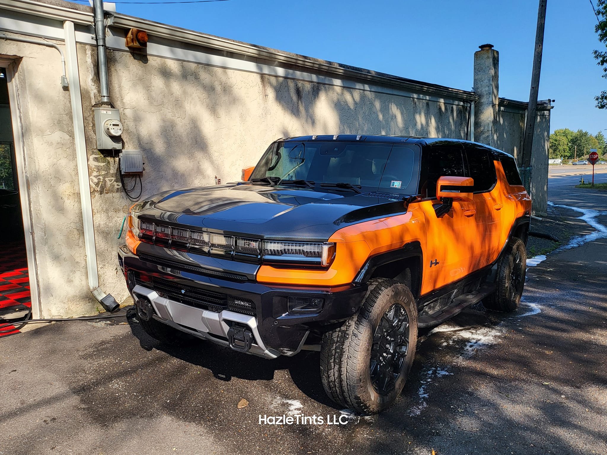 A black and orange SUV parked outside a building with a textured light-colored wall, electrical meters, and a sidewalk. There is a stop sign and some trees in the background.