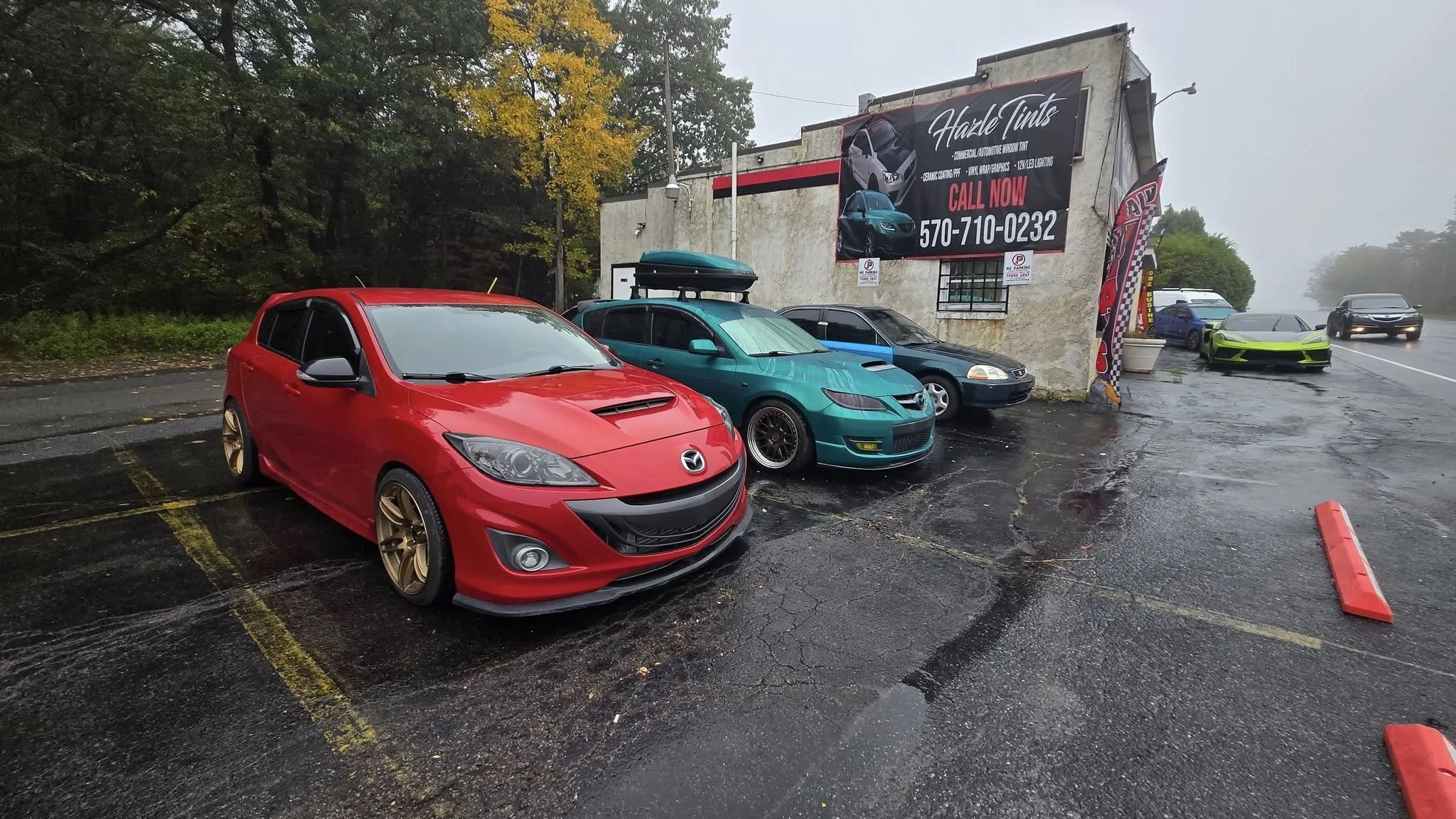 A parking lot with several cars, including a red Mazda with gold rims, a green car with a roof cargo box, and a black sports car, next to a building with a sign for auto tinting services. The ground appears wet, and there are trees in the background.