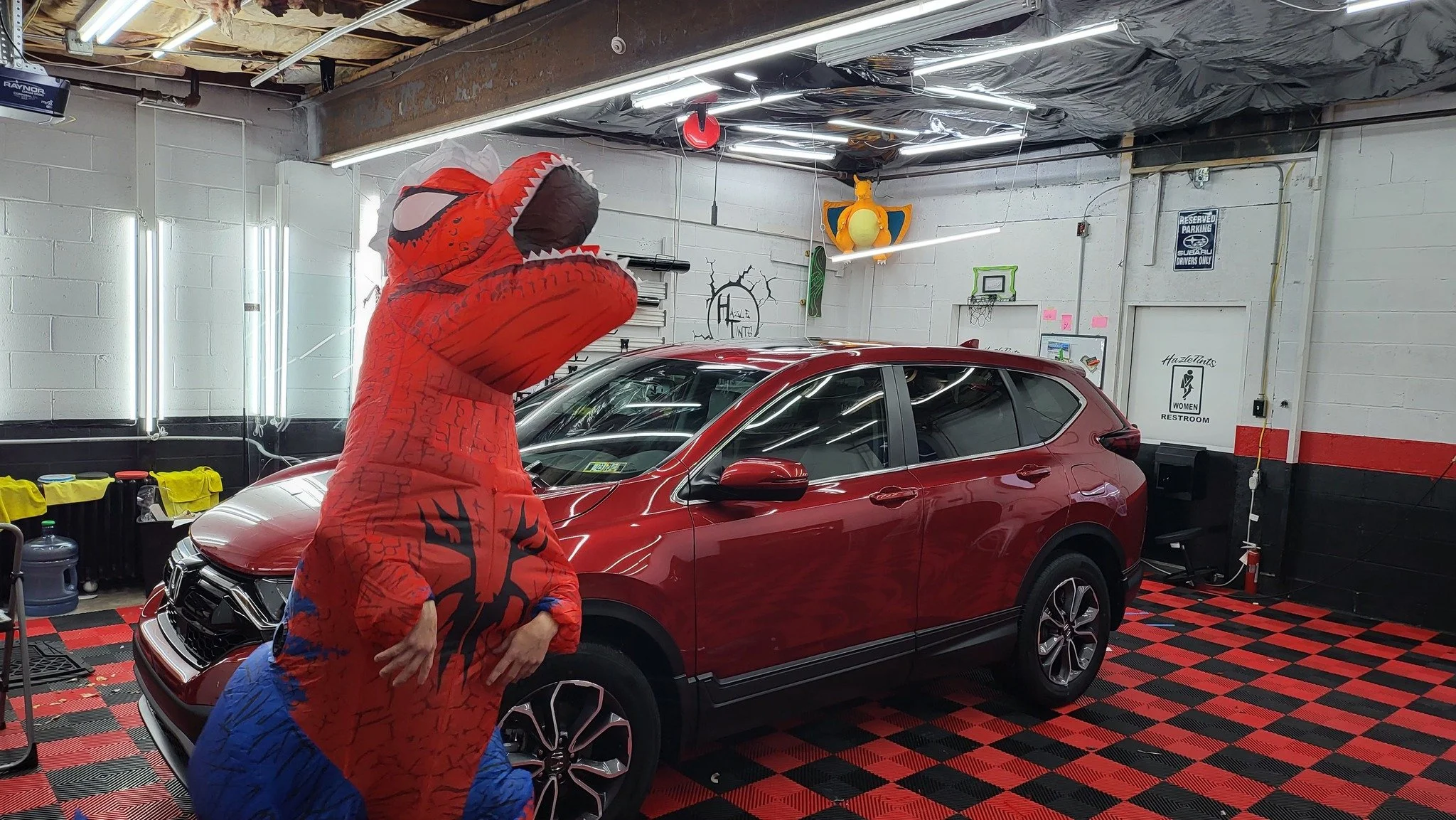 Person in a Spider-Man costume posing next to a red car inside a garage or auto shop.