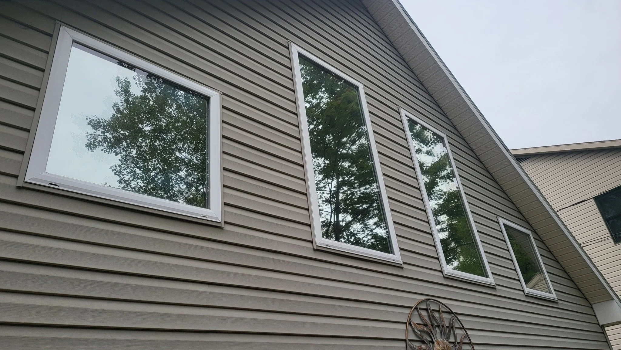 Close-up of the exterior of a house with beige siding, featuring several tall rectangular windows reflecting trees, and a decorative metal sunburst wall art below the windows.