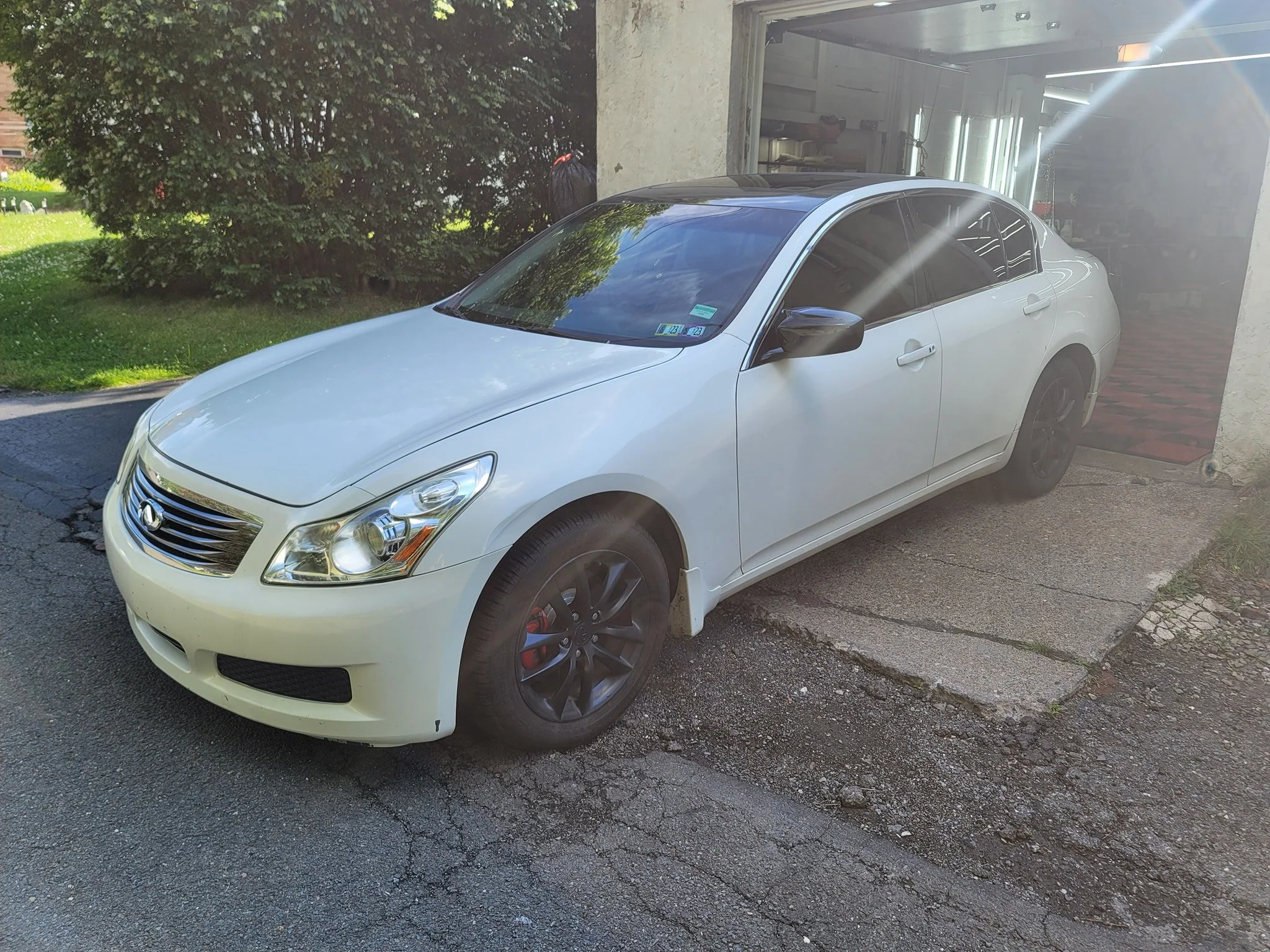 A white sedan parked on a driveway outside a garage with a partially open door. The car has dark tinted windows and black wheels.