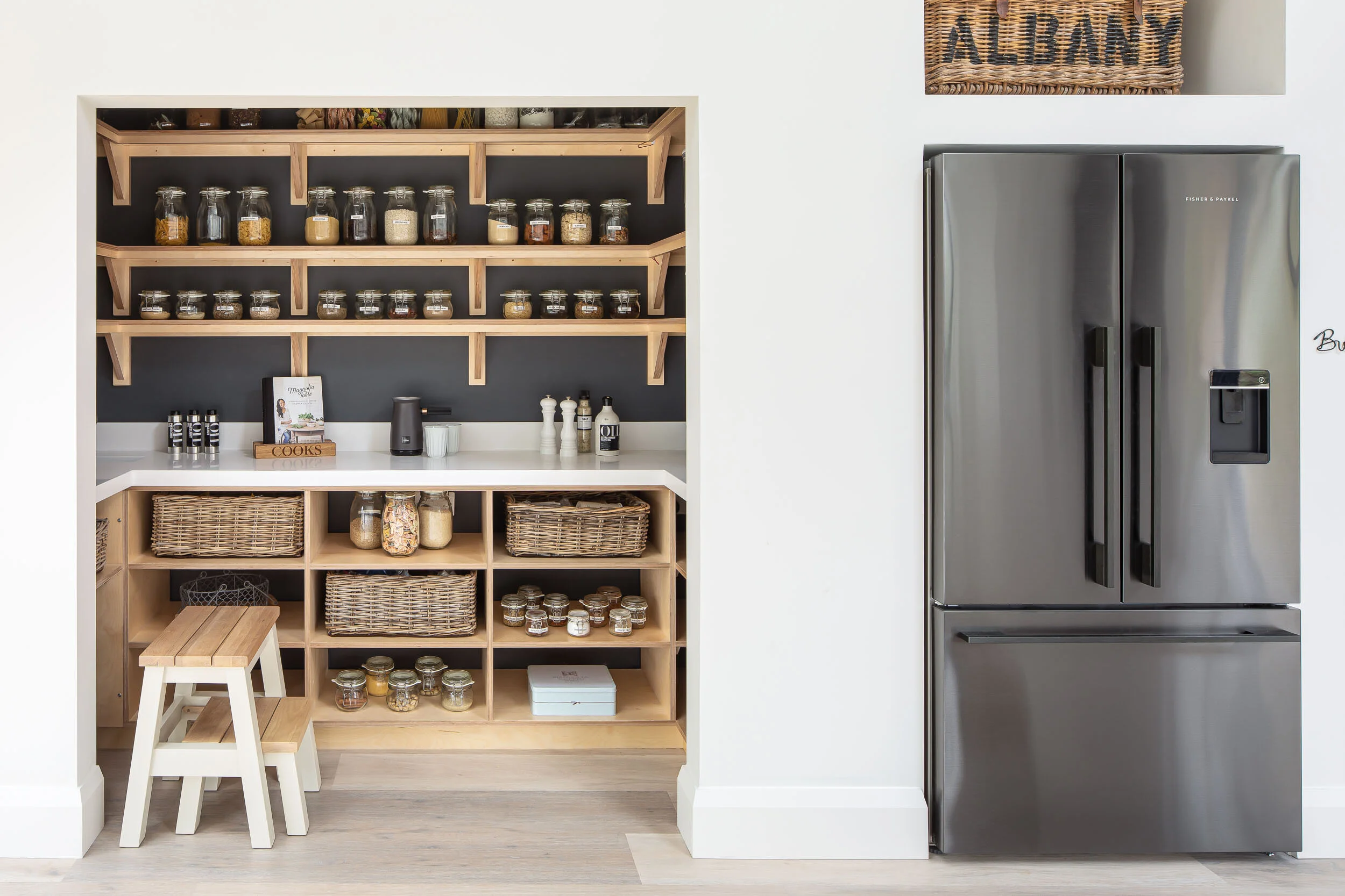 Pantry with open shelving, glass jars filled with dry goods, wicker baskets, and a stainless steel refrigerator on the right. A small wooden step stool is in front of the shelves, and a variety of kitchen items like a cookbook, spice bottles, and a c