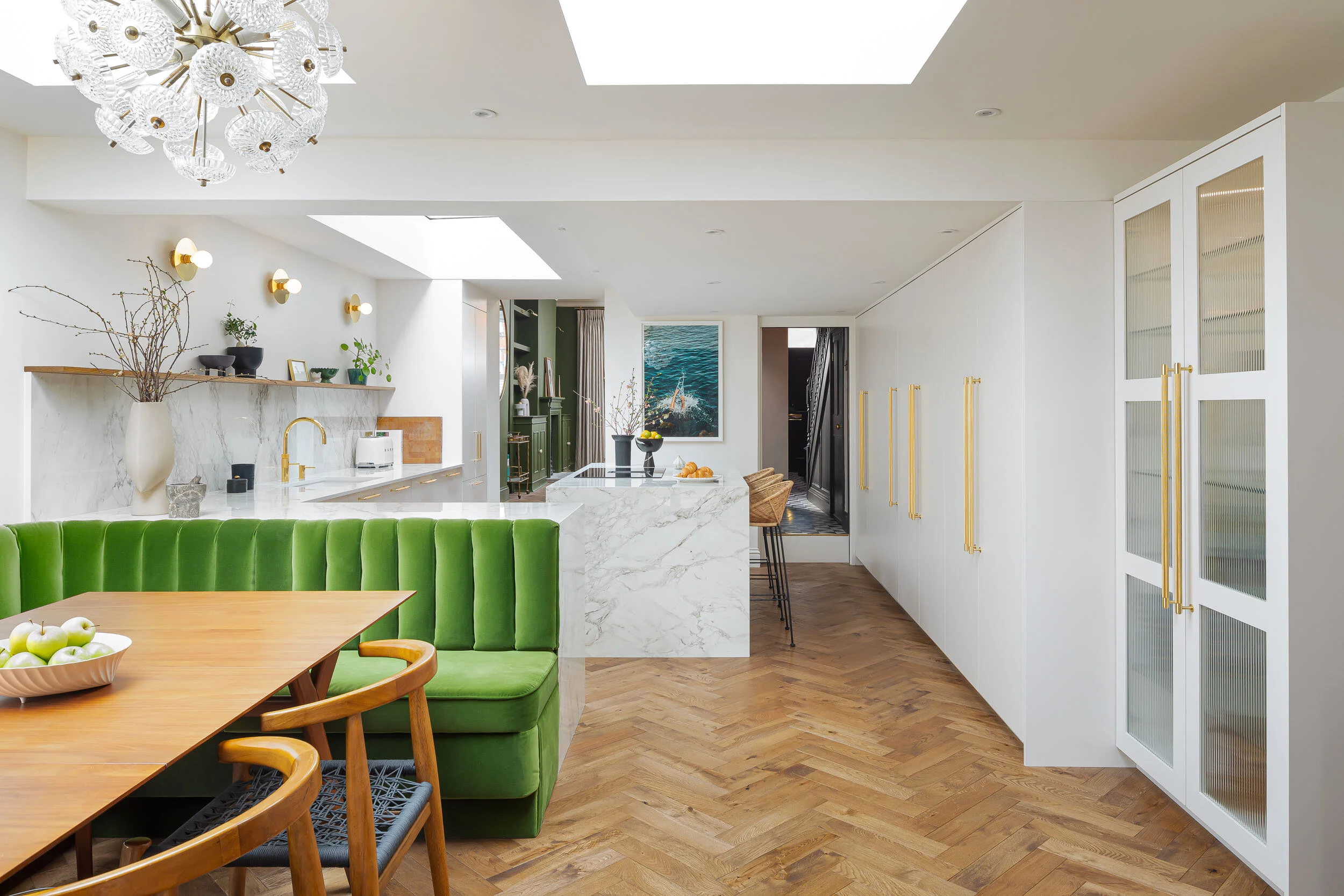 Modern kitchen and dining area with green banquet seating, wooden table, and chairs. Features a marble countertop, gold accents, and natural decor. Overhead lighting fixture and skylights provide ample lighting. Hardwood herringbone floor, contempora