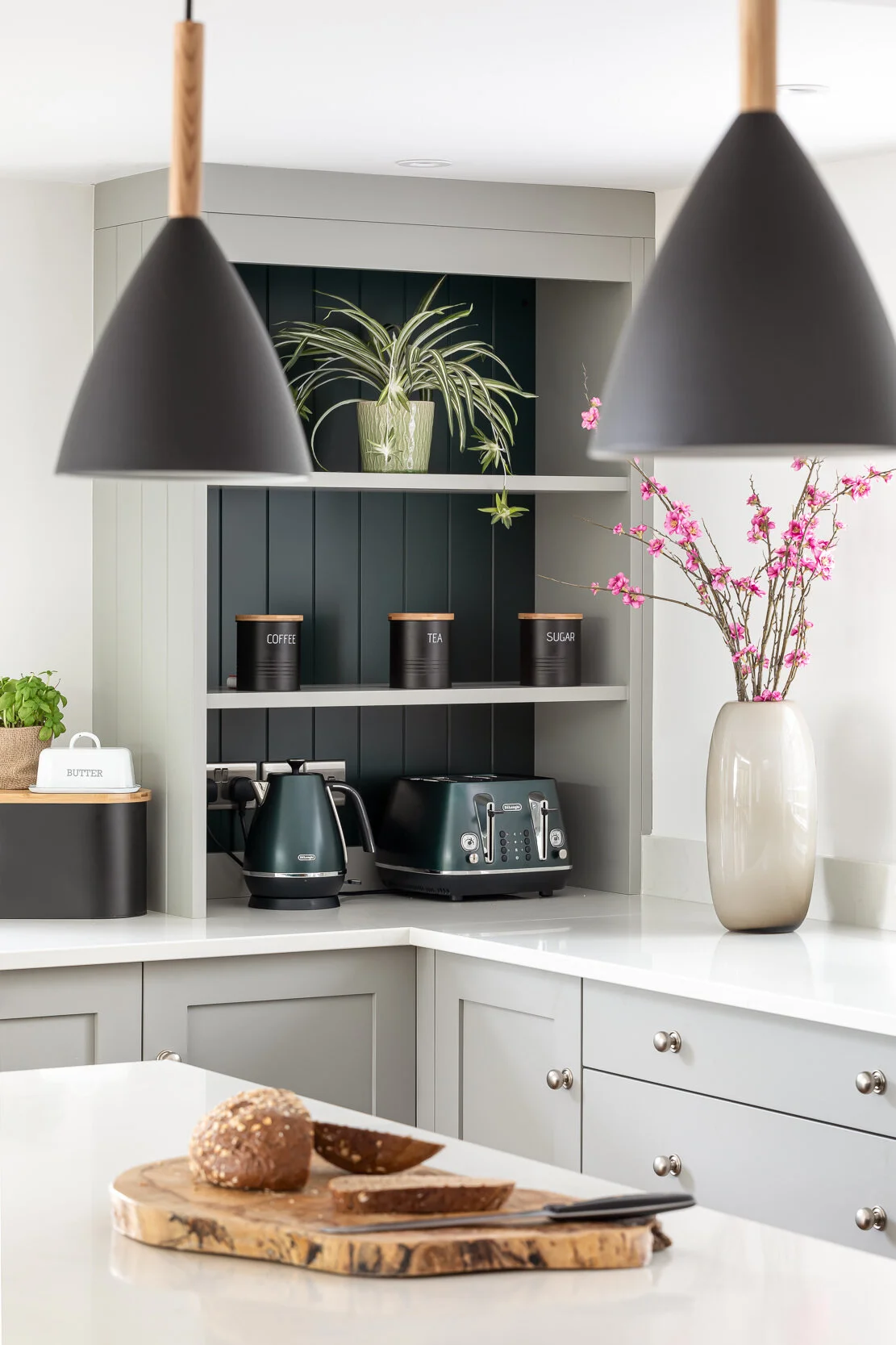 Modern kitchen with green cabinets and countertops, featuring black pendant lights, a vase with pink flowers, a potted plant, black canisters labeled coffee, tea, and sugar, a kettle, toaster, and a wooden cutting board with sliced bread.