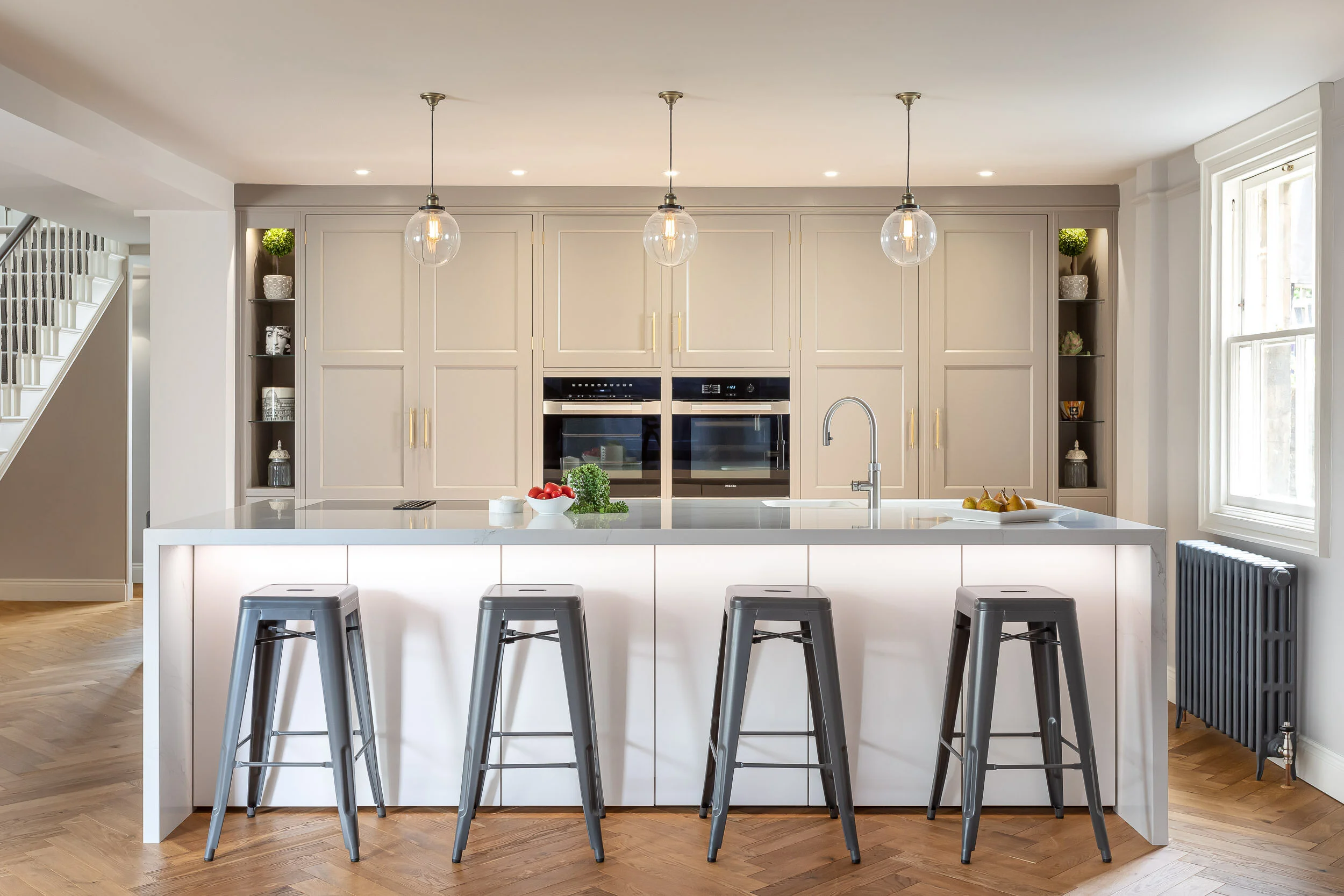 Modern kitchen with white cabinetry, a large island with bar stools, pendant lighting, and built-in ovens. The island features a sleek faucet and decorative fruit. Hardwood flooring and open shelving add warmth.