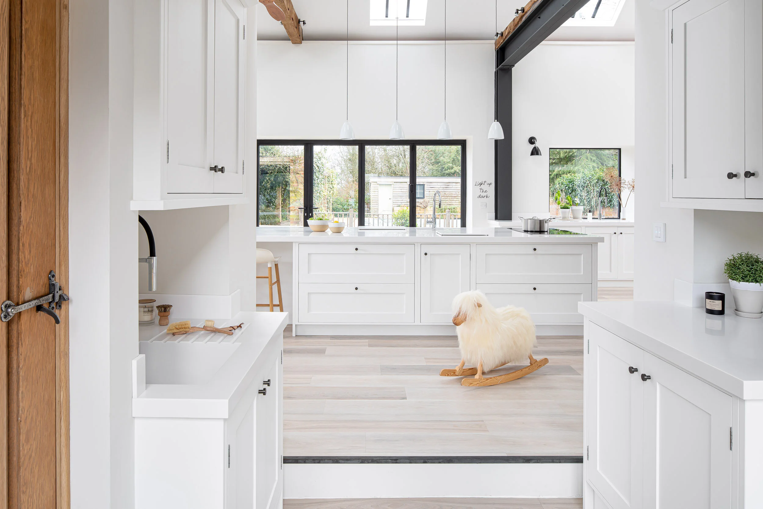 Modern white kitchen with a large island, wooden beams, skylights, and a rocking sheep toy on the floor. The room has large windows, wood flooring, pendant lights, and various decor elements like a small plant and candles.