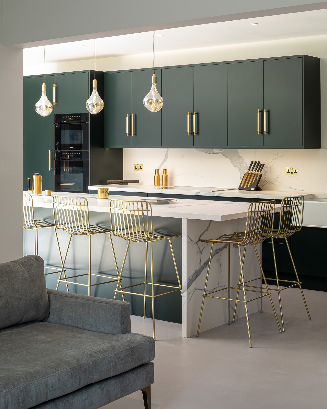 Modern kitchen with green cabinets, white countertops, gold bar stools, and pendant lights. Includes a marble island and a gray sofa in the foreground.