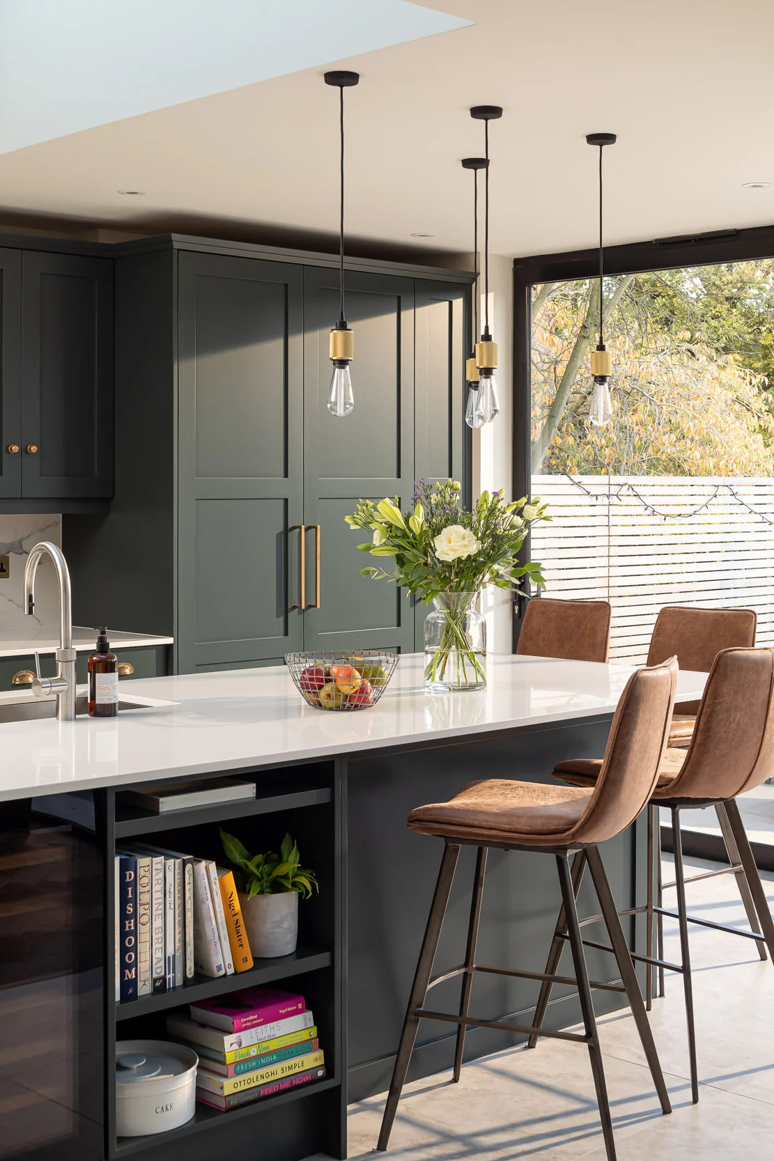 Modern kitchen with dark green cabinets, three pendant lights, and a white countertop featuring a fruit basket and flower vase. Leather bar stools line the island, which has open shelving with books and a plant. A large window provides natural light 