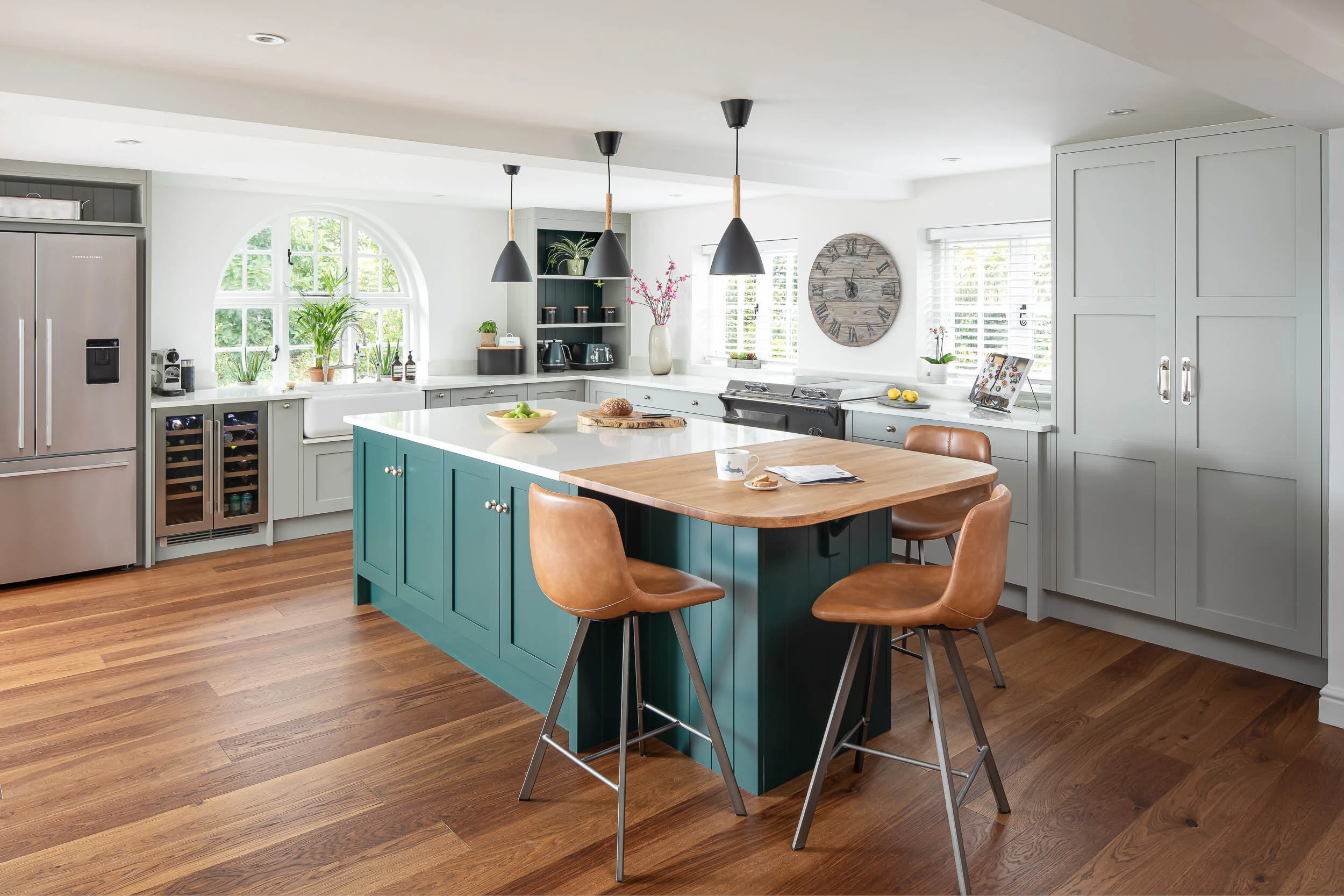 Modern kitchen with teal island, brown leather stools, stainless steel refrigerator, wine cooler, wooden flooring, pendant lights, and large window with greenery.