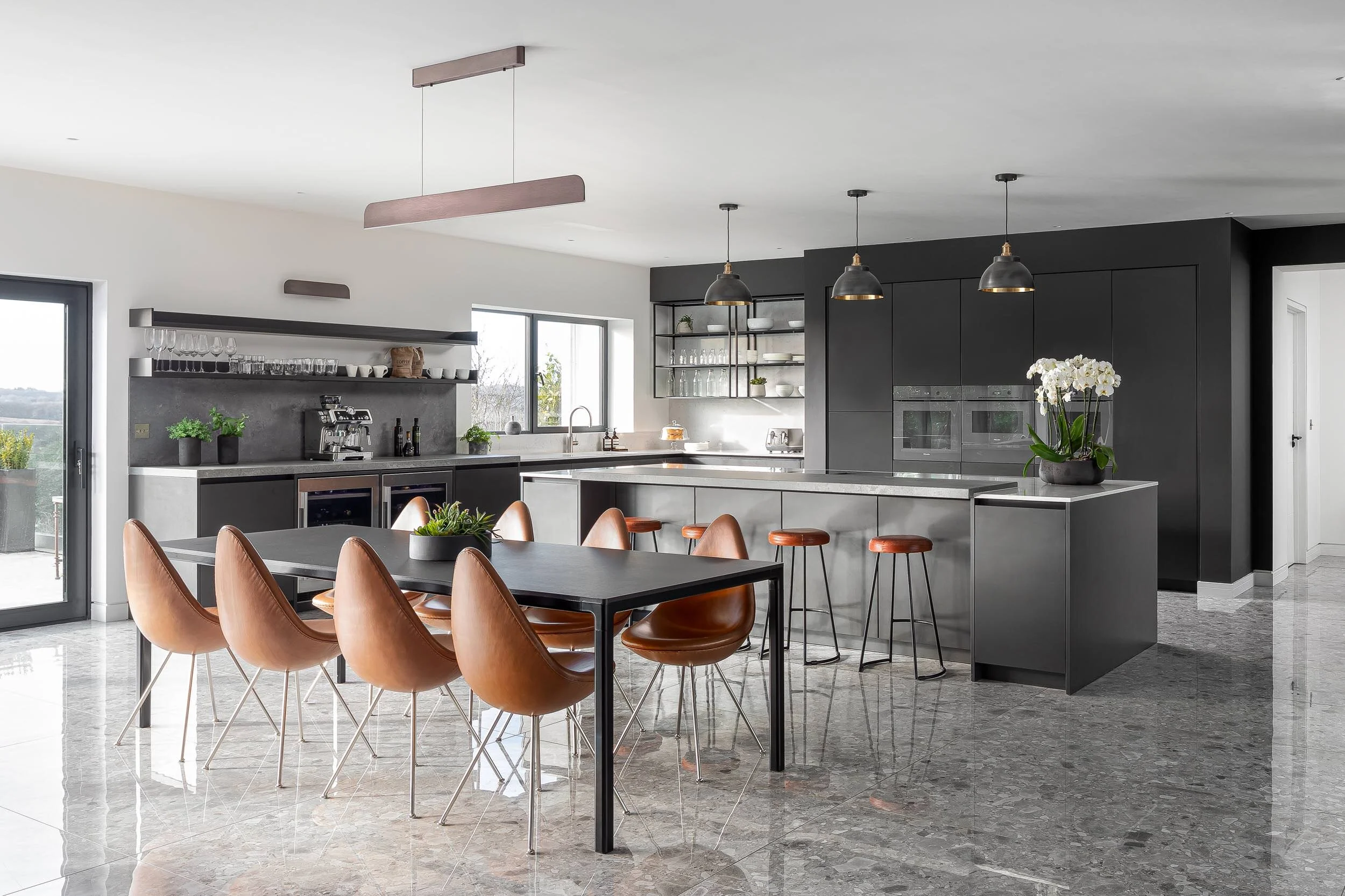 Modern kitchen with black cabinetry, stainless steel appliances, kitchen island with bar stools, dining table with brown leather chairs, pendant lighting, and large windows.