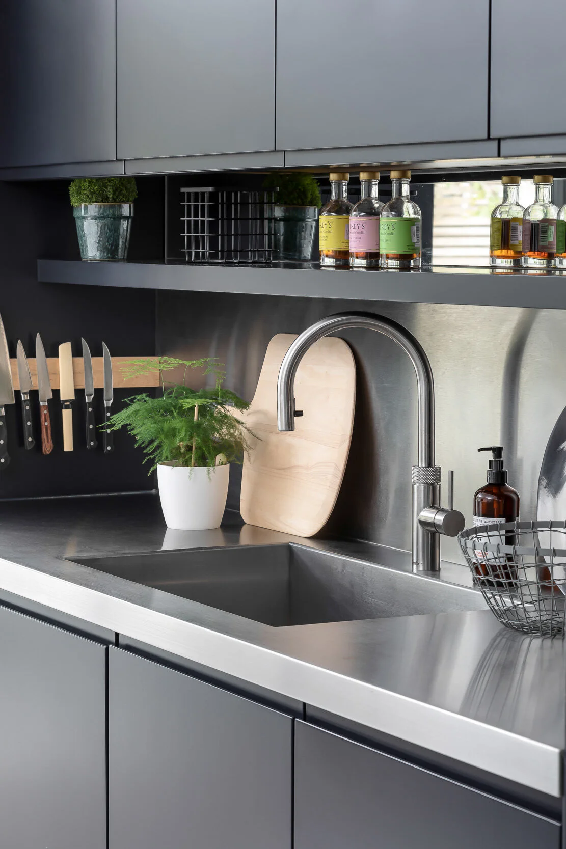 Modern kitchen with stainless steel countertop, faucet, and sink. A magnetic knife strip holds knives, and a small potted plant is beside a cutting board. Glass bottles and items on shelves in background.