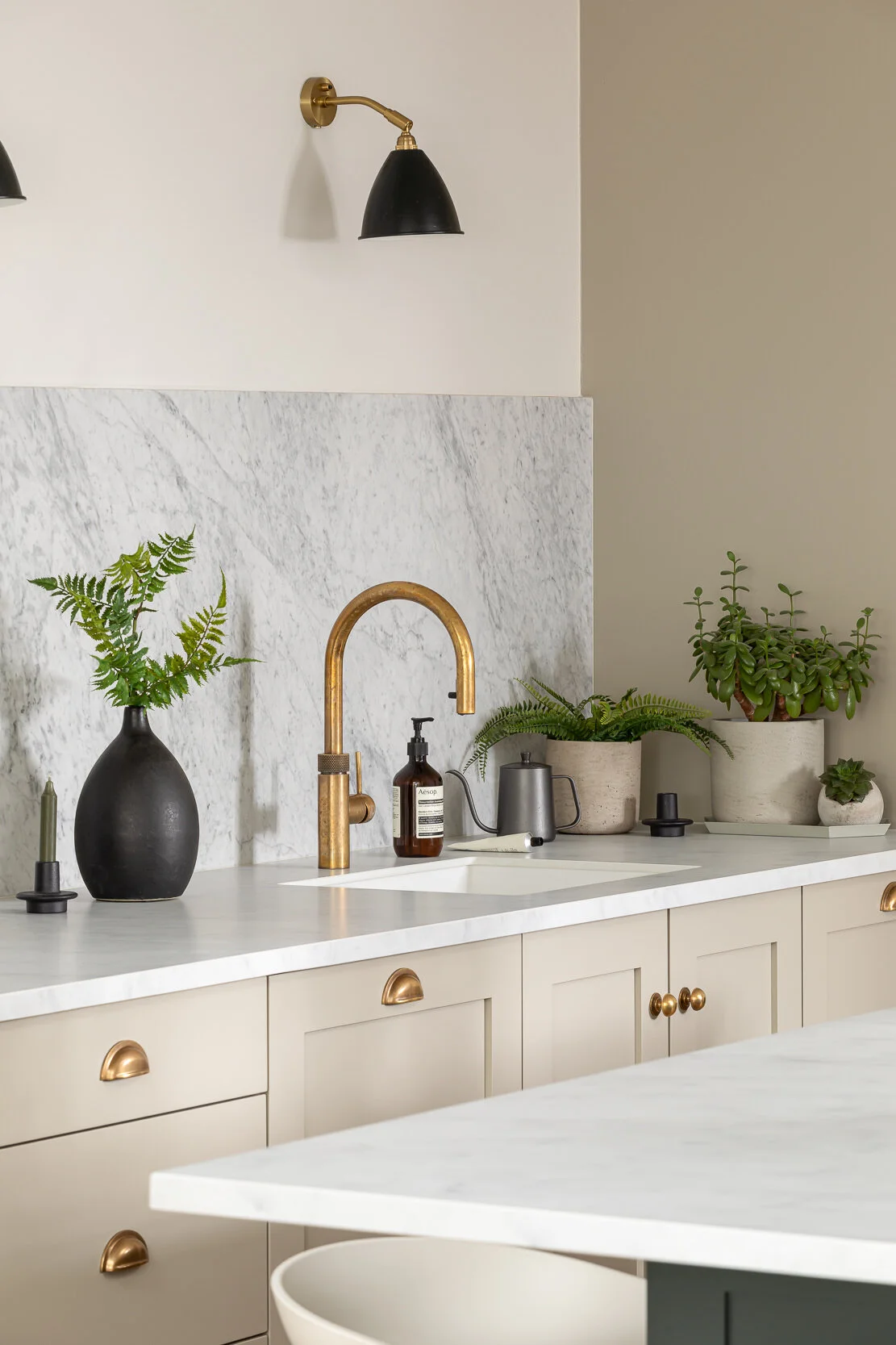 Modern kitchen with a marble backsplash, brass faucet, black wall-mounted lamps, and houseplants on the countertop.