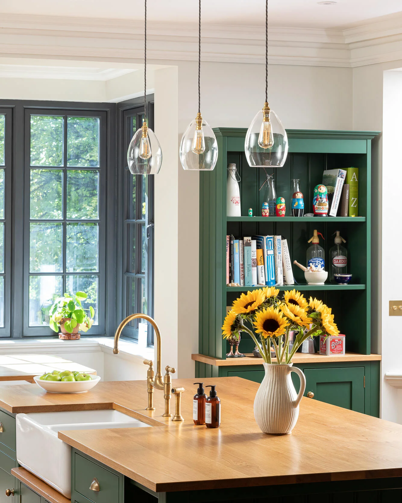 Modern kitchen with green cabinetry, wooden countertops, and pendant lighting. A vase of sunflowers sits on the counter near a brass faucet and soap dispensers. A bowl of green apples is by a window, and shelves display books and decorative items.