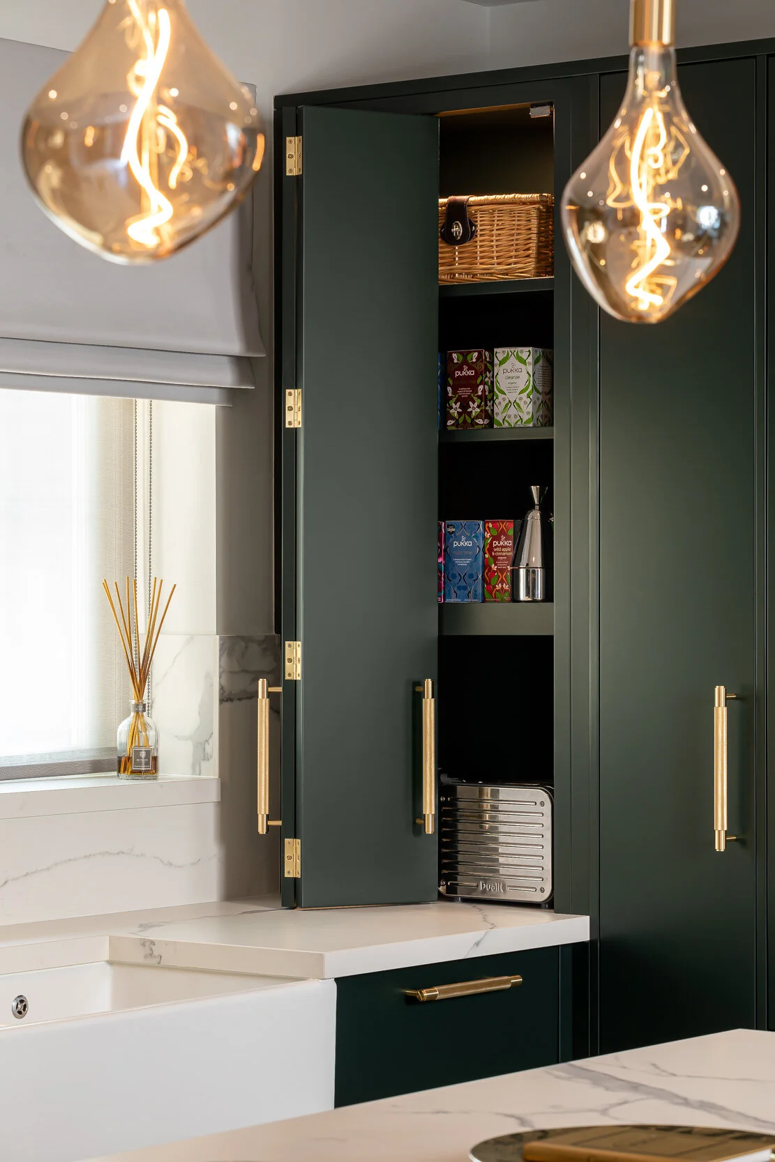 Modern kitchen with dark green cabinets, open cupboard showing tea boxes and a teapot, marble countertops, gold hardware, and decorative light bulbs.
