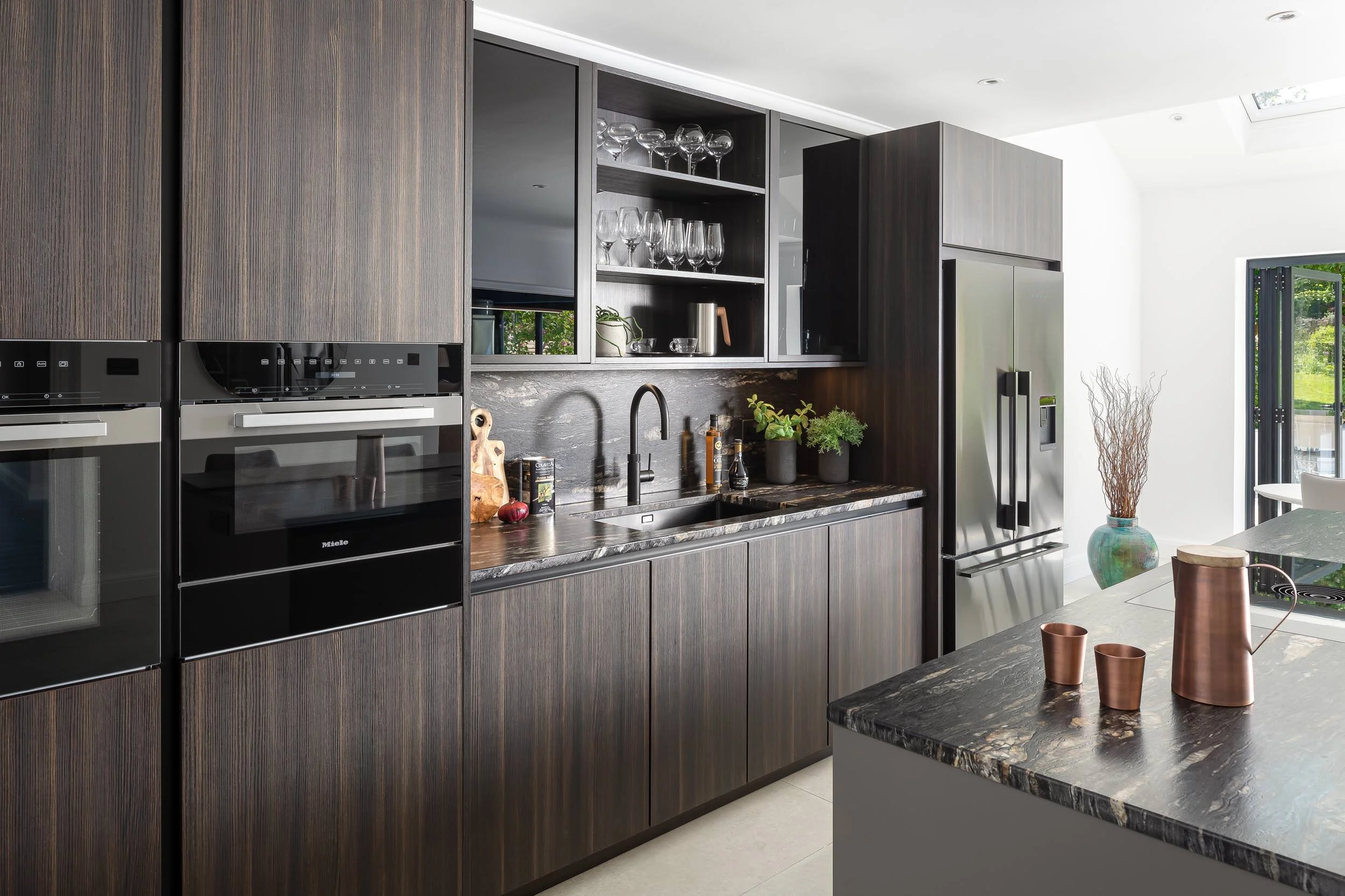 Modern kitchen with dark wooden cabinetry, stainless steel appliances including a double oven and refrigerator, black marble countertops, a built-in sink with a black faucet, open shelving with glassware, and decorative plants on the counter.