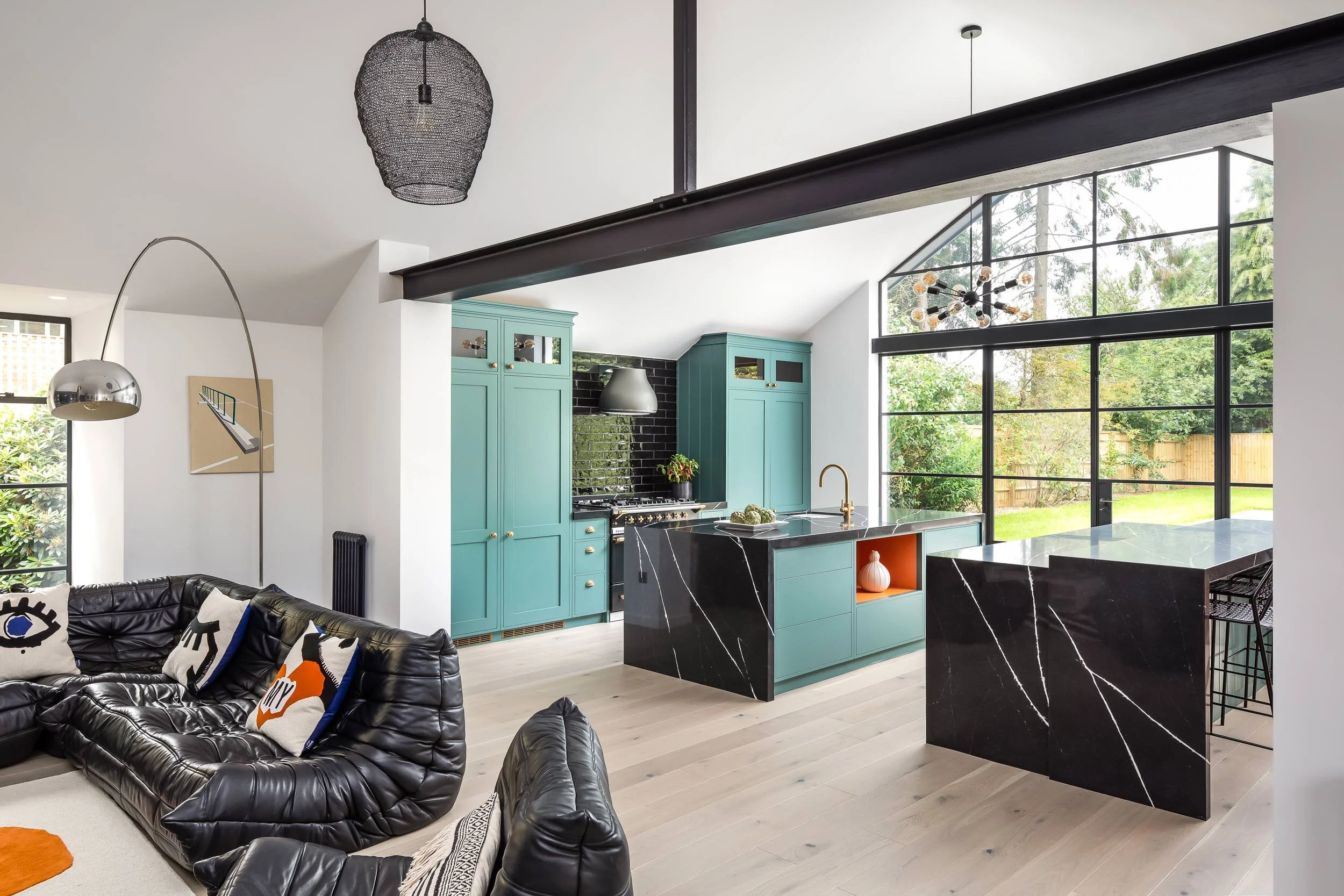 Modern open-plan living room and kitchen with stylish furnishings, including a black sectional sofa, colorful cushions, teal cabinets, black kitchen island, and large windows overlooking a garden.