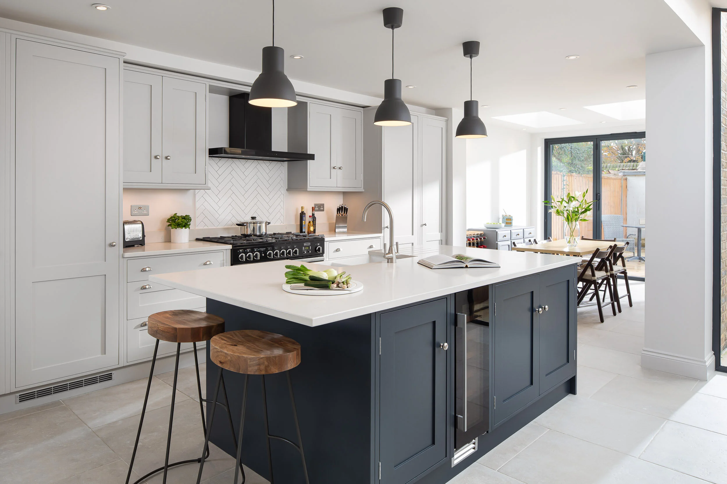 Modern kitchen with a large island, pendant lighting, and wooden stools. White cabinets, black appliances, and a tiled backsplash are present. A dining area with a table and chairs is visible near large windows.