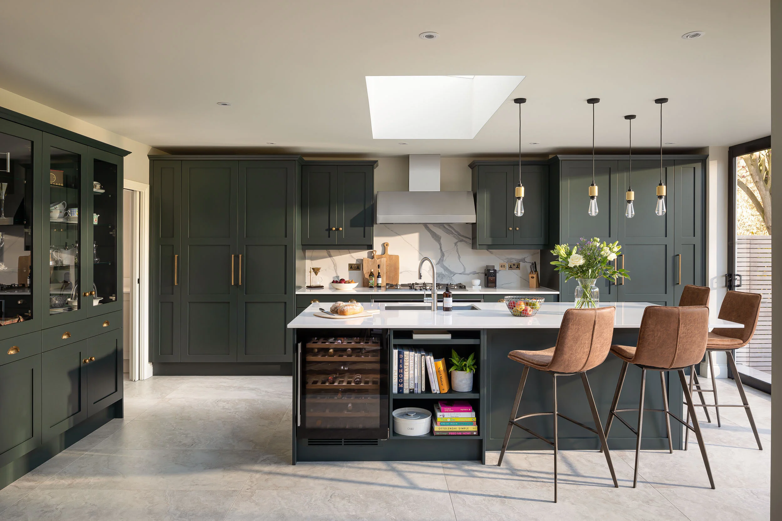 Modern kitchen with dark green cabinetry, a marble backsplash, and a large island with brown stools. The island features built-in shelving, a wine fridge, and a white countertop. Hanging pendant lights and a skylight illuminate the space, which also 