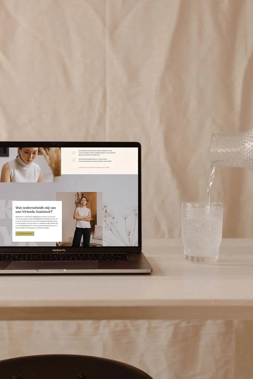 Een open MacBook Pro laptop op een lichte houten tafel met een glas water dat wordt ingeschonken. Achter de tafel hangt een beige gordijn.