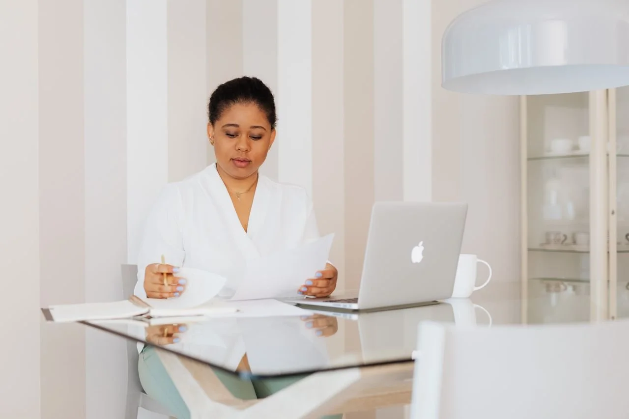 Cosmetische kliniek ondernemer leest documenten aan een glazen bureau met laptop in een minimalistisch, licht interieur