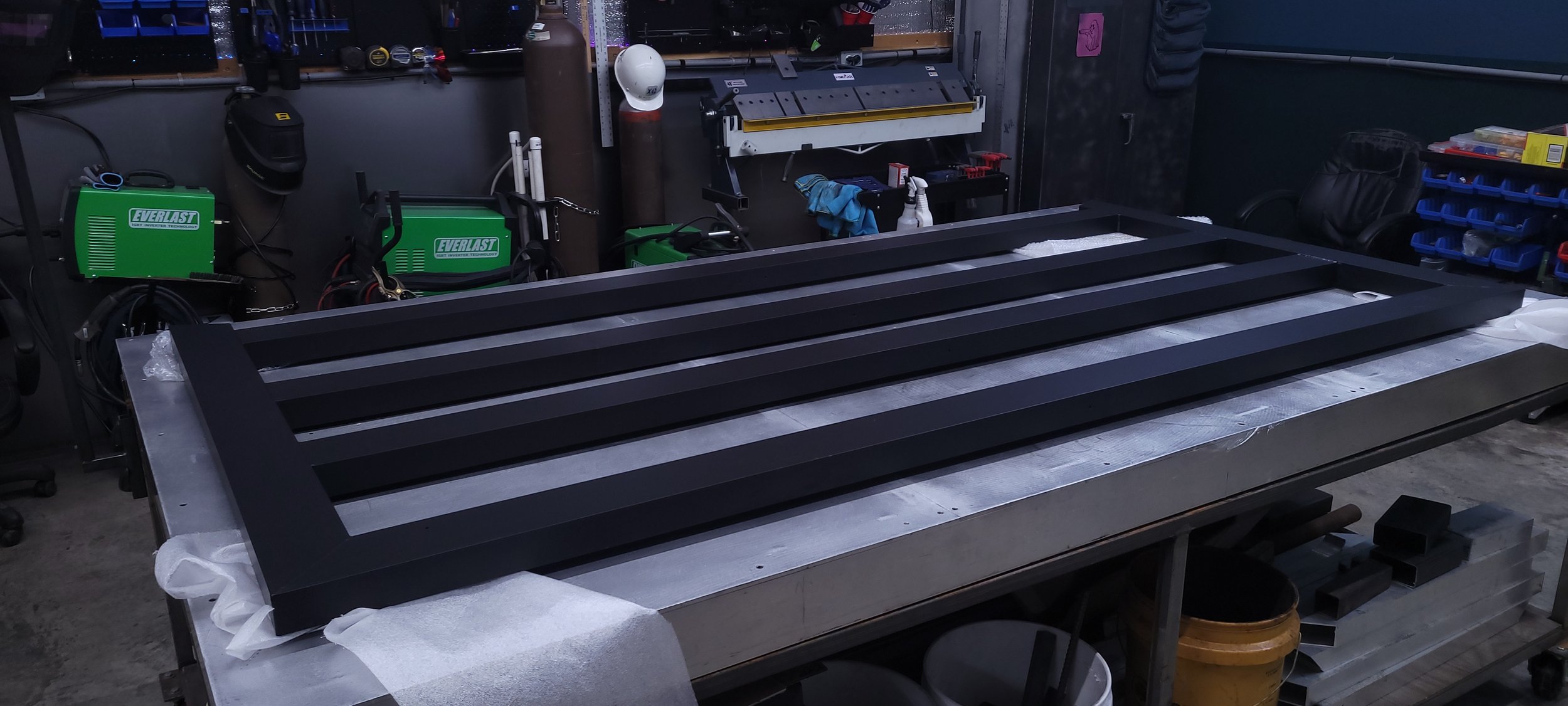 A workshop workbench with a large black metal frame, possibly a gate or a panel, in process of being assembled, with tools and supplies around.