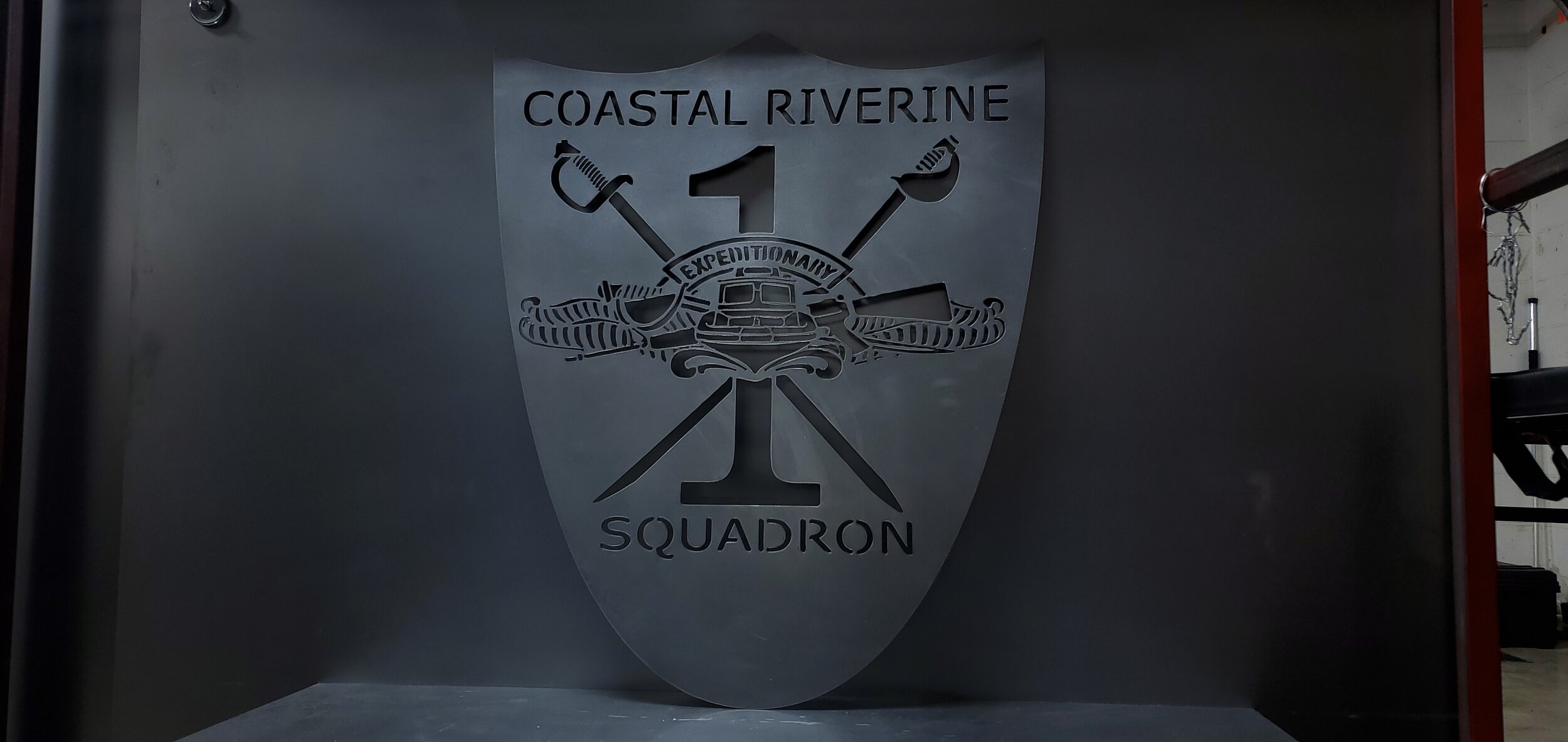 A metallic shield with the words 'Coastal Riverine 1 Squadron' and a central emblem depicting a boat, with crossed knives and tools, and armed with a knife, fork, and spoon.