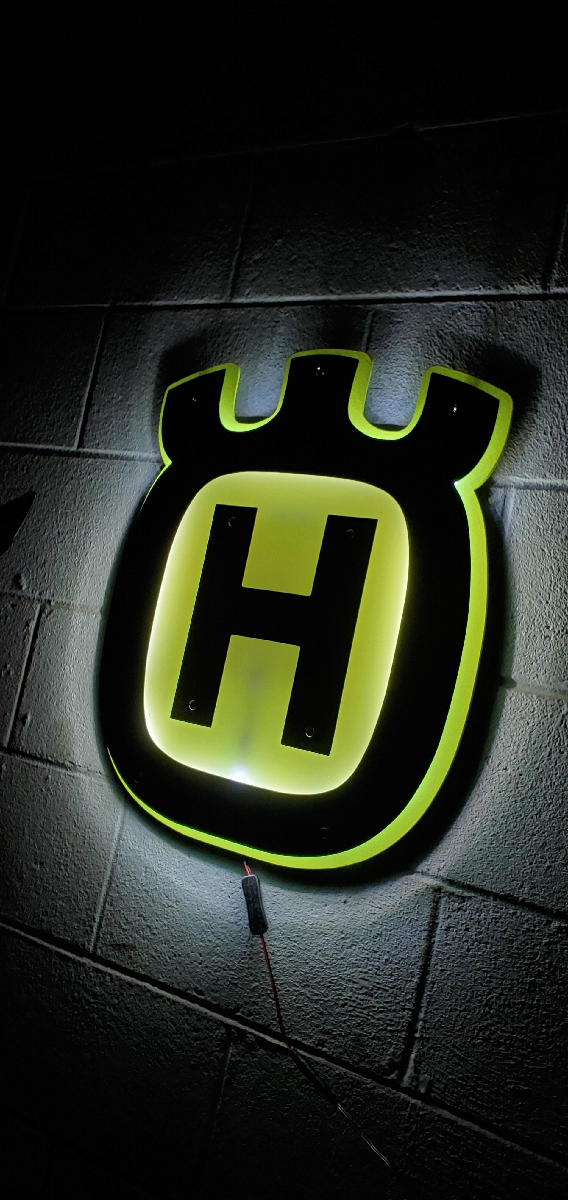 Bright illuminated sign with a black and yellow shield-shaped logo featuring a large letter H in the center, mounted on a textured wall.
