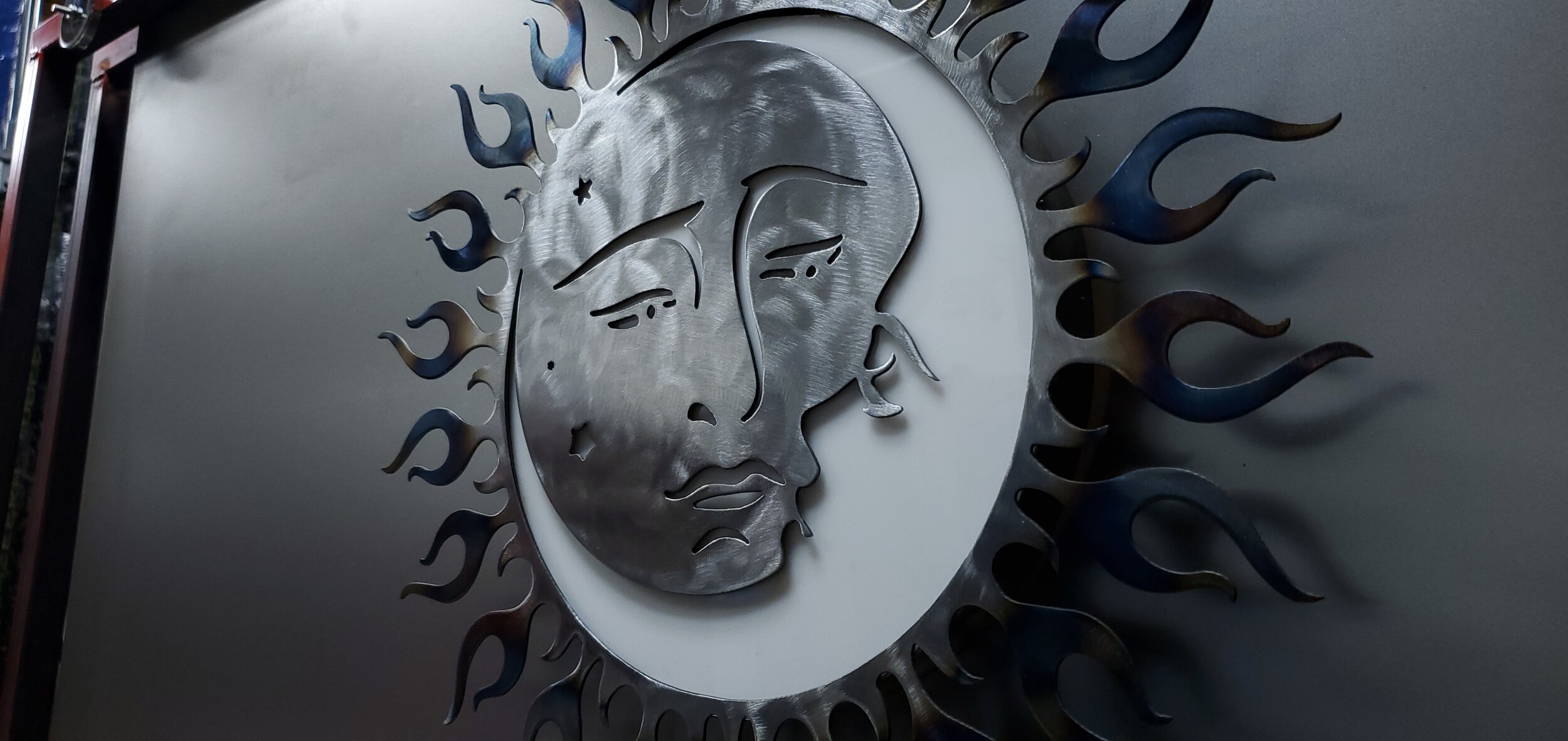 Metal art piece depicting a stylized human face with sun-like rays surrounding it, mounted on a wall.