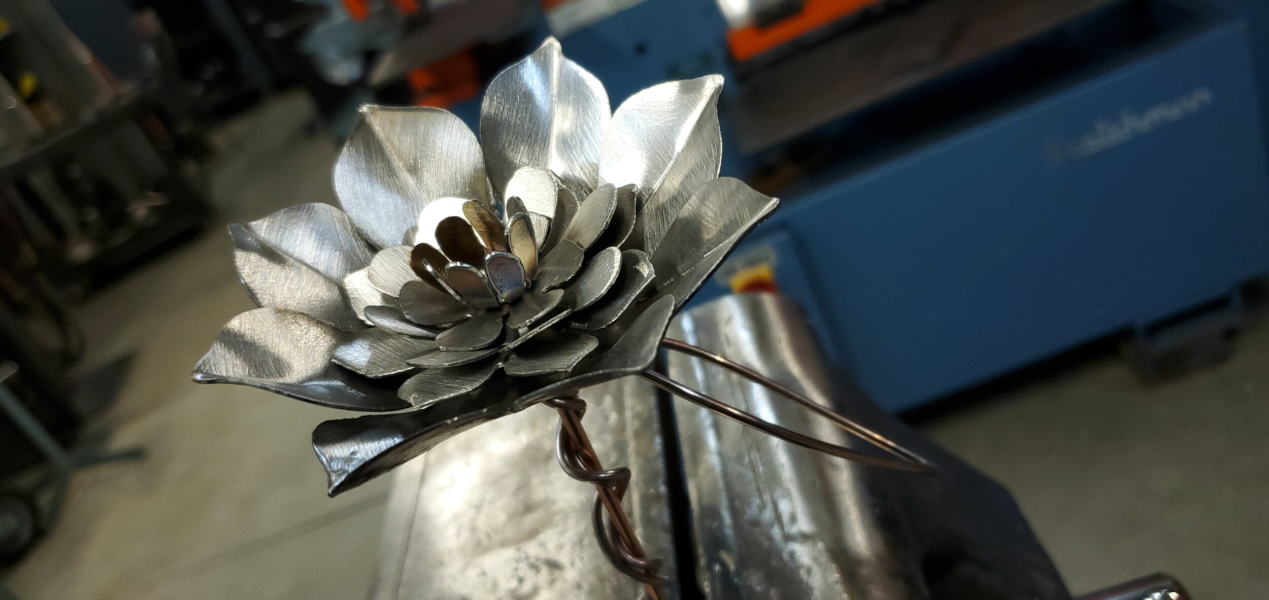A metal flower sculpture with layered petals, crafted from silver-colored metal, attached to a twisted wire stem.