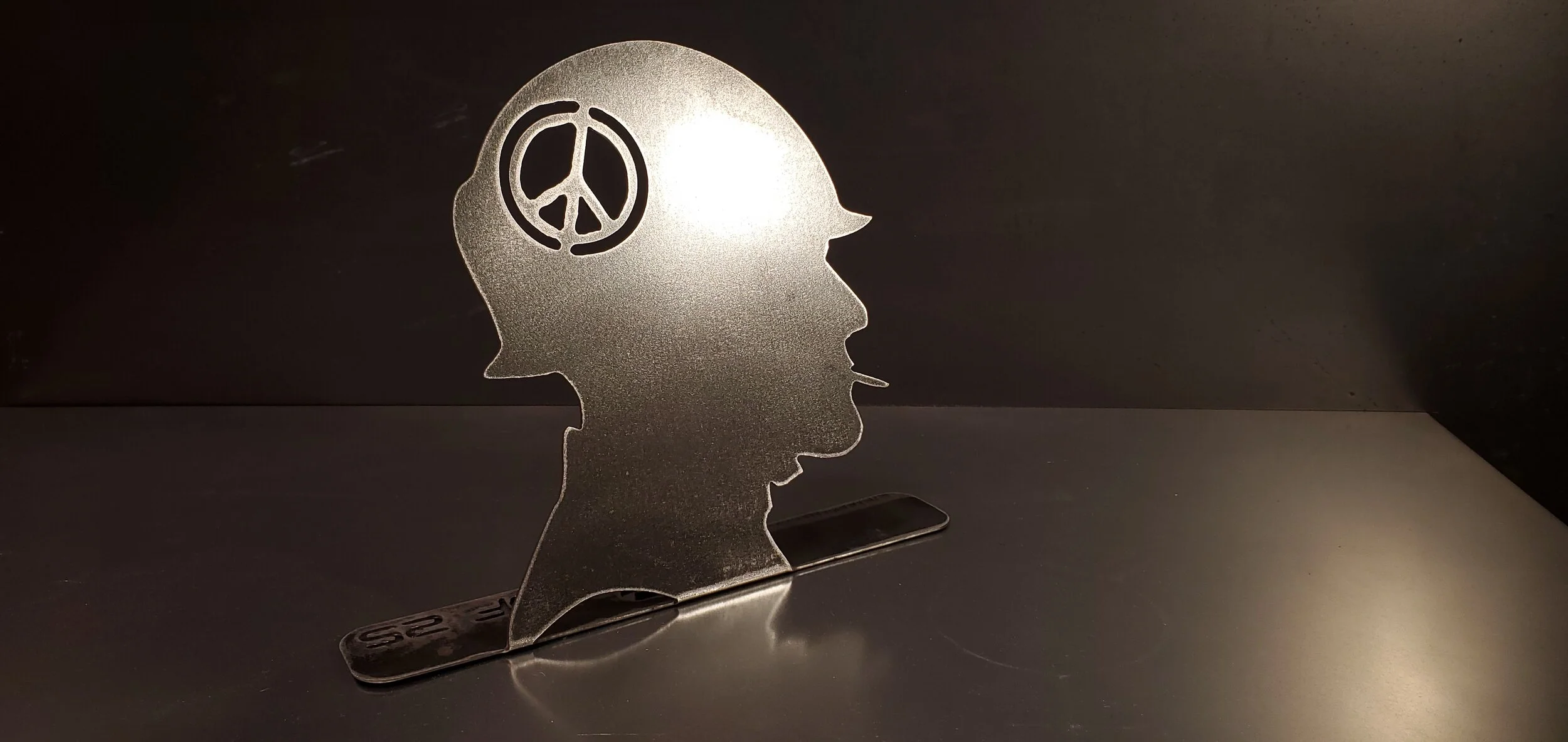 Silhouette of a human head with a peace symbol in the brain area, made of metallic material, standing on a counter with a dark background.