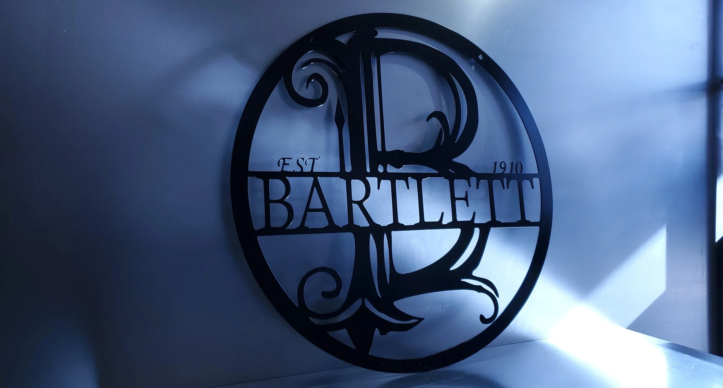 Black metal circular sign with the name 'BARTLETT' and the year 1910, mounted on a wall with dramatic shadows.