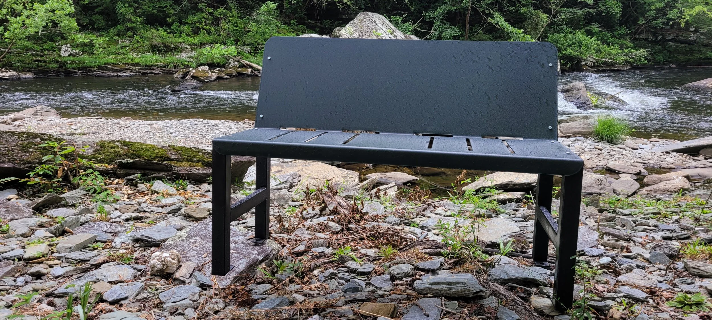 A black metal bench on rocky ground by a river, with green foliage and rocks in the background.
