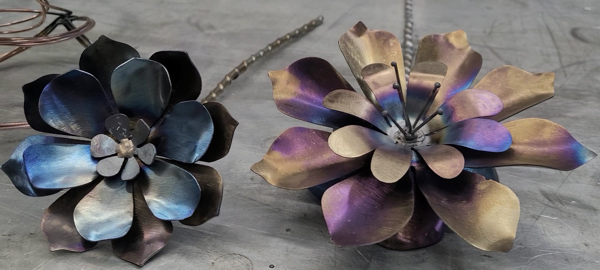 Two metal flower sculptures, one with blue and black petals and the other with gold and purple hues, resting on a metallic surface.