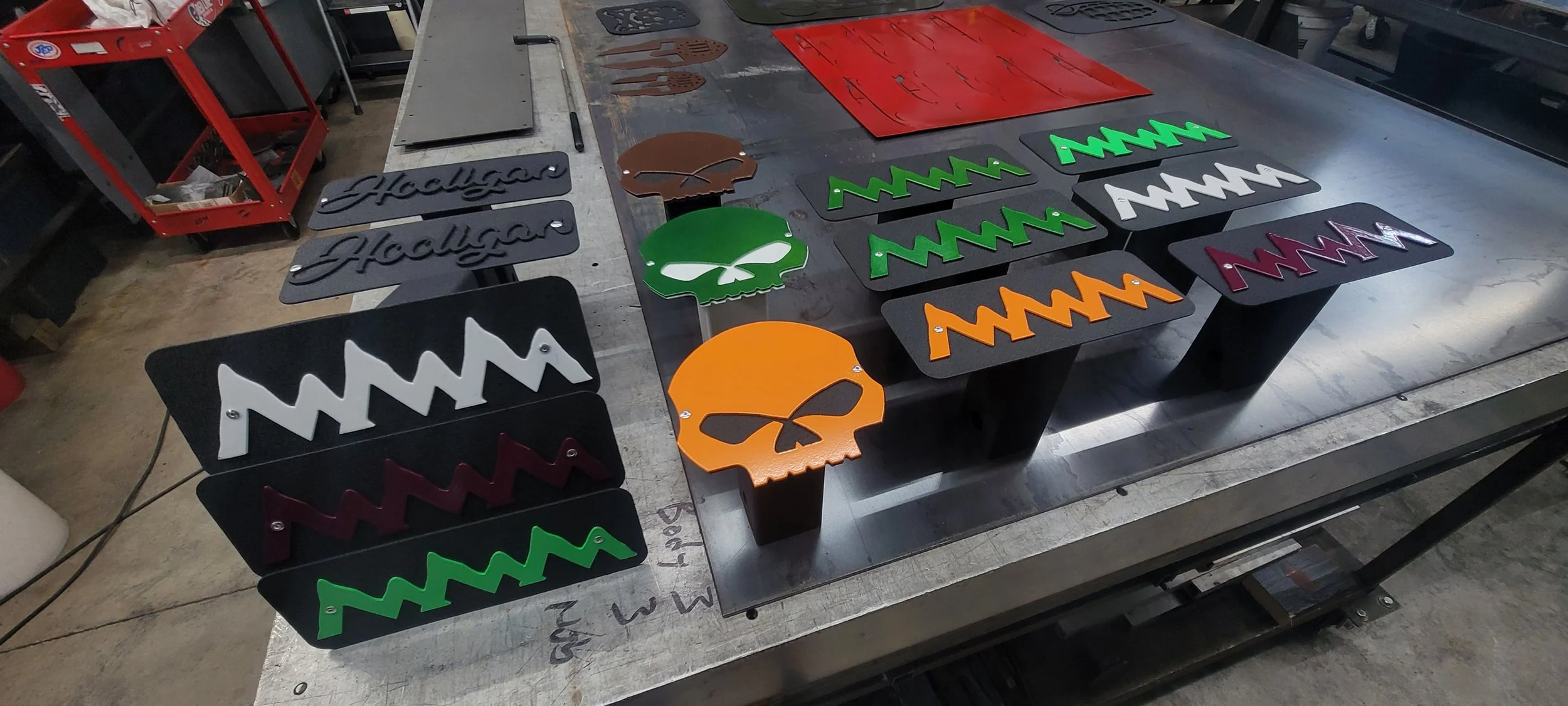 Metal signs with various designs, including words and skull graphics, laid out on a worktable in a workshop. Made by Turn 2 Customz