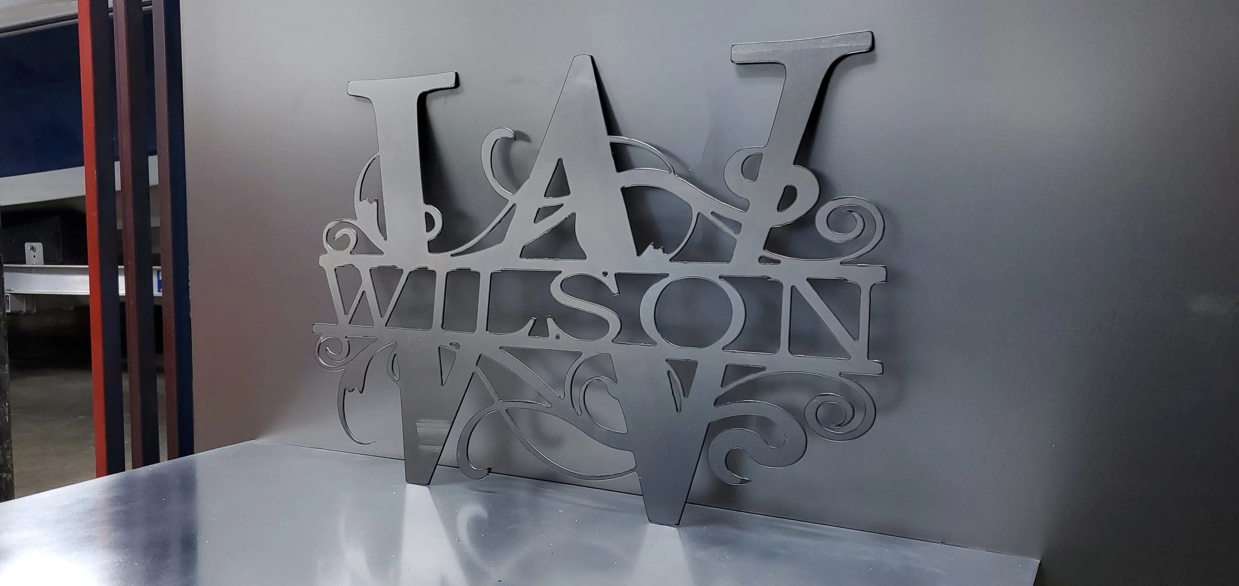 Metal wall art with the words 'LA WILSON' in a decorative, cut-out design, placed on a reflective surface against a plain wall.