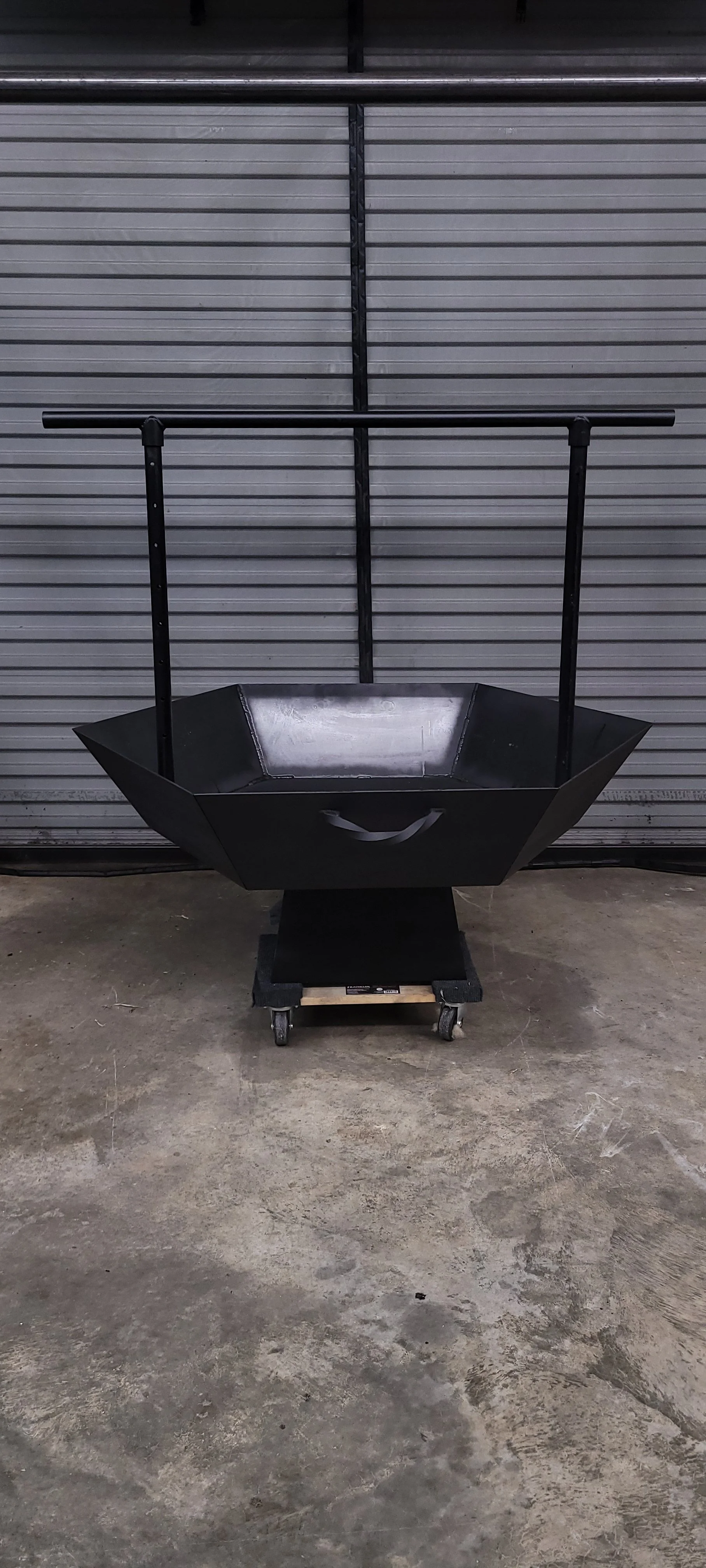 A black metal fire pit with a hexagonal shape on caster wheels in front of a closed, gray metal garage door. Medium custom steel fire pit designed and fabricated by Turn 2 Customz in East Tennessee