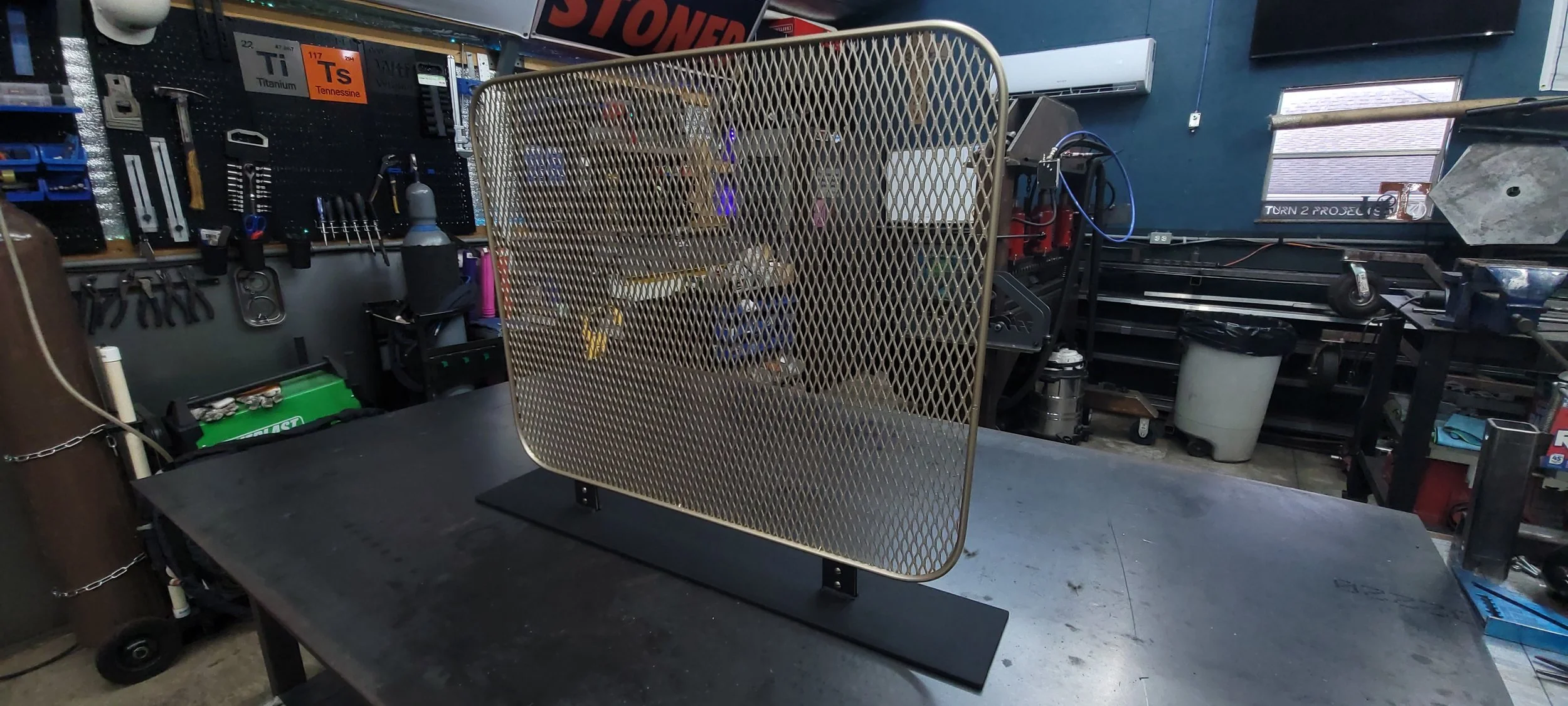 A workshop with a metal mesh safety shield on a workbench, surrounded by tools, equipment, and a pegboard with hand tools on the wall.